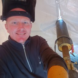 mart spaw - Autoportret spawacza w masce, z uśmiechem, na tle białego namiotu i spawanej rury. Widoczne ślady spawania na rurze i narzędzia w tle.