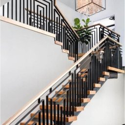 mart spaw - Nowoczesne schody z czarną, metalową balustradą o geometrycznym wzorze, drewnianymi stopniami i podświetleniem. W tle widoczna roślina doniczkowa i dekoracyjna lampa.