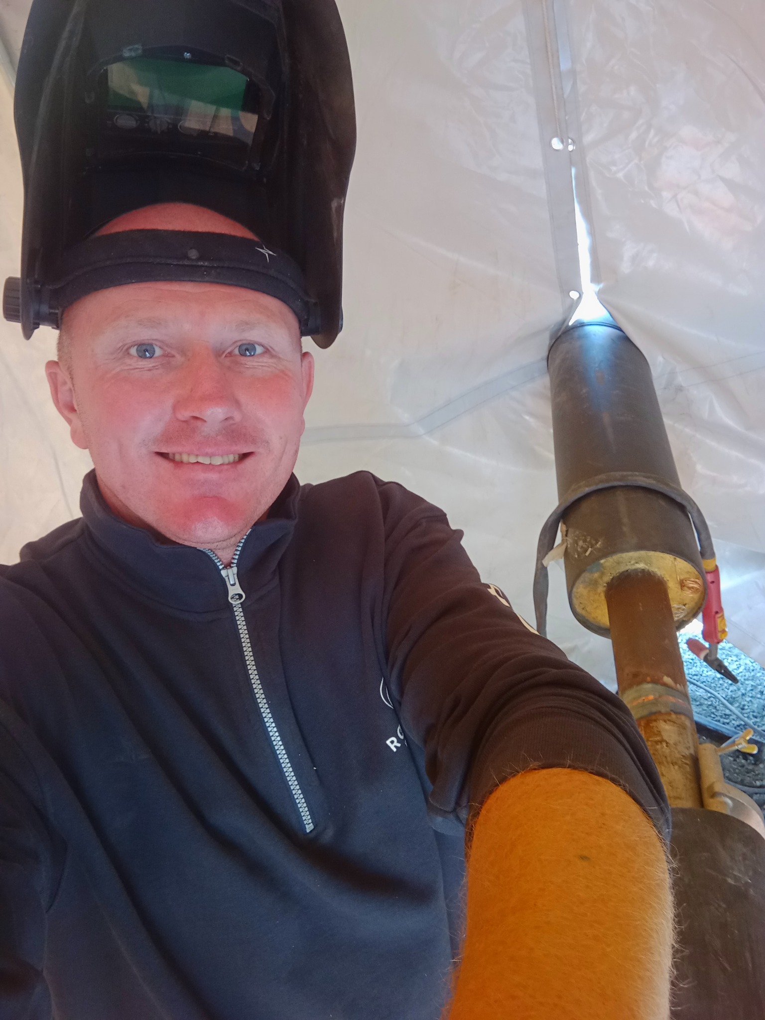 Autoportret spawacza w masce, z uśmiechem, na tle białego namiotu i spawanej rury. Widoczne ślady spawania na rurze i narzędzia w tle.