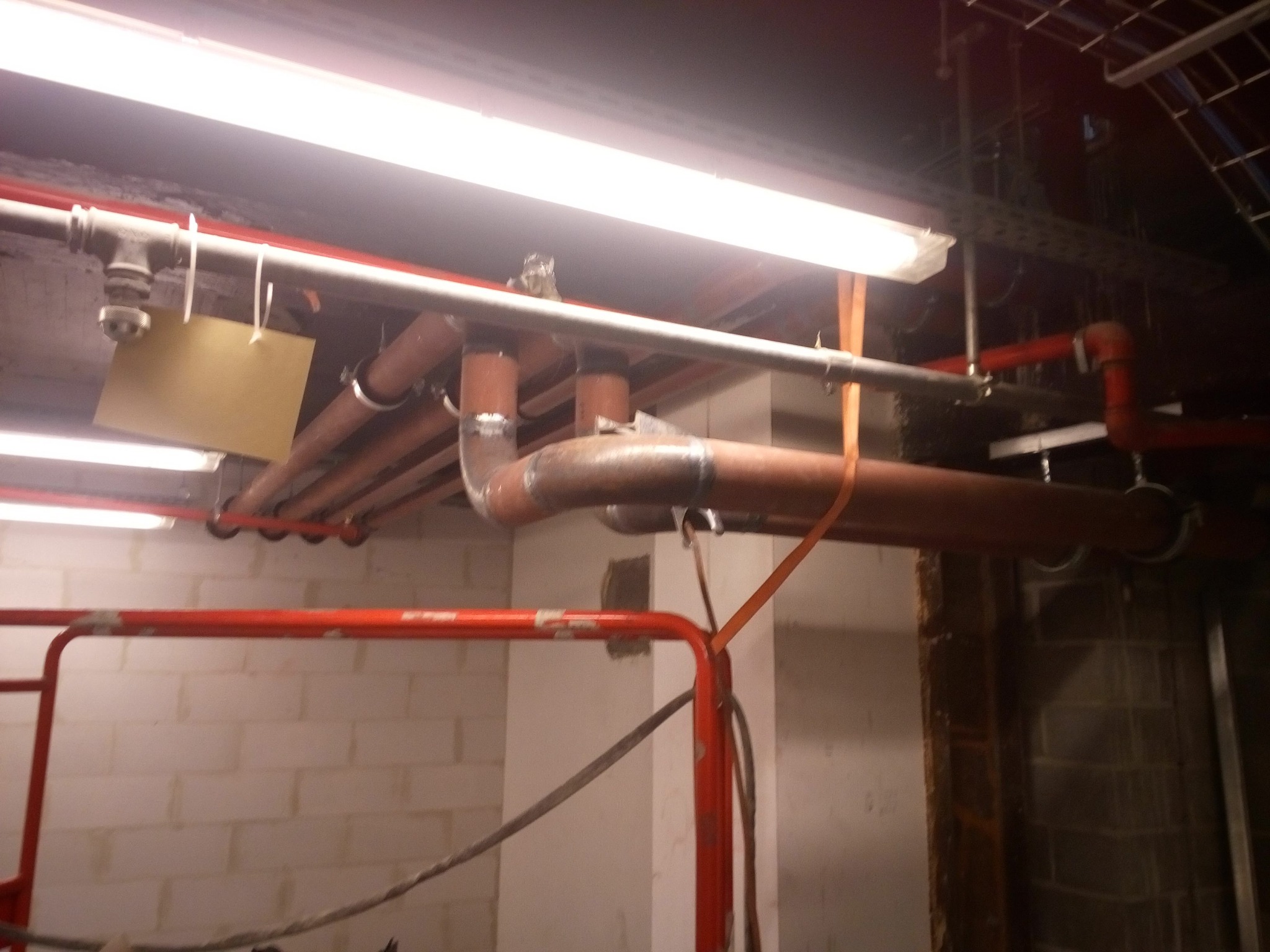 Instalacja rur w budynku w Woli Murowanej. Widoczne rury miedziane, stalowe i elementy mocujące pod sufitem. Prace spawalnicze w trakcie realizacji.