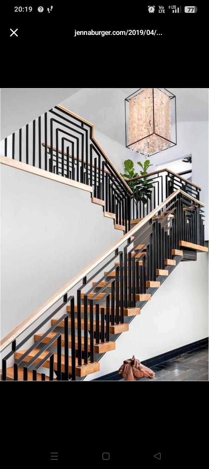 Nowoczesne schody z czarną, metalową balustradą o geometrycznym wzorze, drewnianymi stopniami i podświetleniem. W tle widoczna roślina doniczkowa i dekoracyjna lampa.