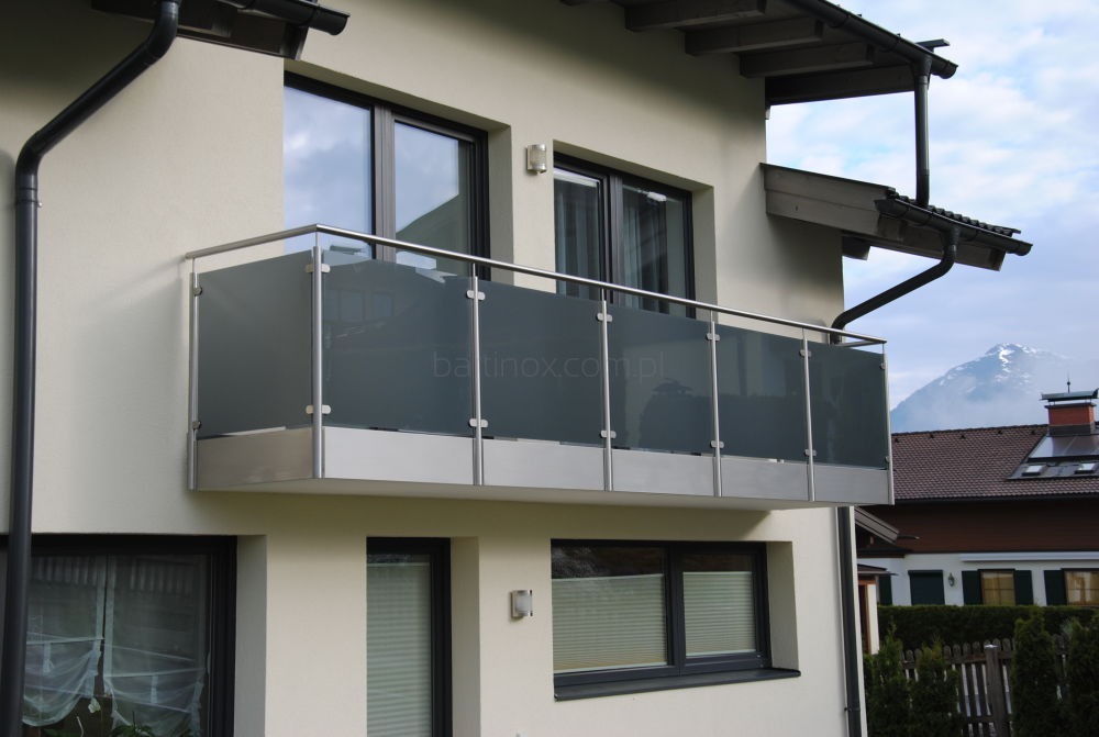 Szklana balustrada balkonowa z metalowymi elementami na tle jasnej elewacji budynku i górskiego krajobrazu w tle. Nowoczesny design z widocznym adresem strony.