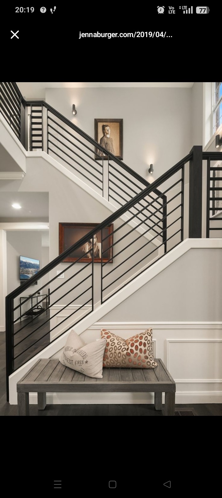 Nowoczesne schody z czarną metalową balustradą, drewniane stopnie, jasne ściany z obrazami i ławka z poduszkami u dołu. Minimalistyczny design.