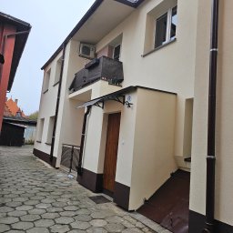 PHU ROBÓT BUDOWLANYCH A. PALUCH - Jasny budynek z ocieploną elewacją, brązowe rynny i podmurówka, widok z wąskiej uliczki wyłożonej kostką.