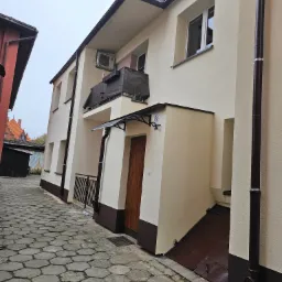 Jasny budynek z ocieploną elewacją, brązowe rynny i podmurówka, widok z wąskiej uliczki wyłożonej kostką.