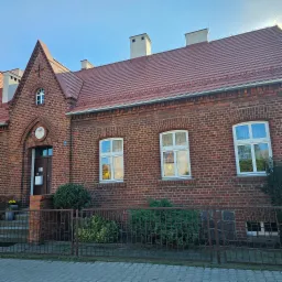 Dwukondygnacyjny budynek z czerwonej cegły z ciemnoczerwoną dachówką, widoczne trzy kominy, okna z białymi ramami i ciemne drzwi wejściowe