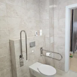 Beżowa łazienka z wiszącą toaletą, uchwytami dla niepełnosprawnych i płytkami imitującymi marmur, widoczny dozownik zapachu i zapas papieru toaletowego.