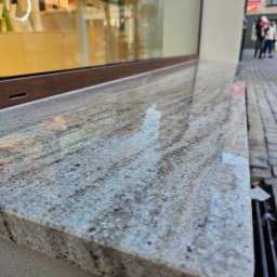 PHU ROBÓT BUDOWLANYCH A. PALUCH - Granitowy parapet zewnętrzny z widoczną fakturą kamienia, zamontowany przy oknie budynku. W tle fragment chodnika i przechodnie.