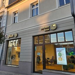 PHU ROBÓT BUDOWLANYCH A. PALUCH - Fasada budynku z szyldami 'plus' i 'polsat box' nad wejściem. Widoczne okna i drzwi wejściowe. Wnętrze biura widoczne przez szybę.