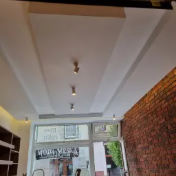 Nowoczesny sufit podwieszany z oświetleniem punktowym w lokalu usługowym, widoczna ceglana ściana i okno wystawowe z nazwą sklepu.