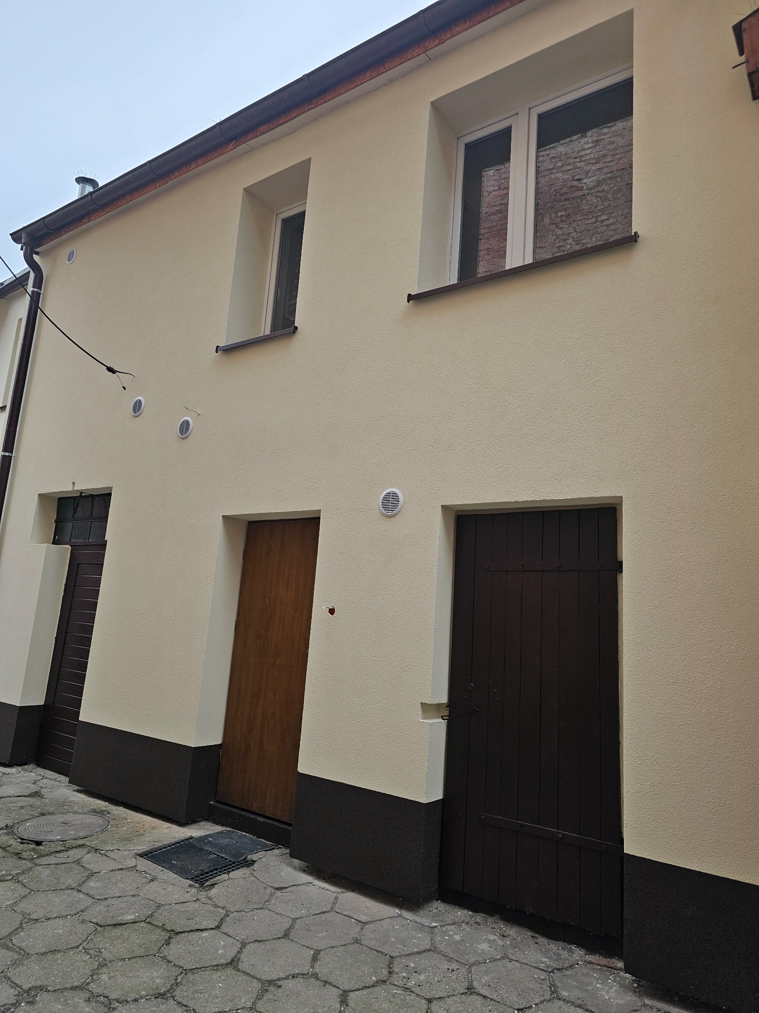 Odnowiona elewacja budynku z kremowym tynkiem, brązowym cokołem i trzema parami drzwi i okien, widok z dołu na szary chodnik.
