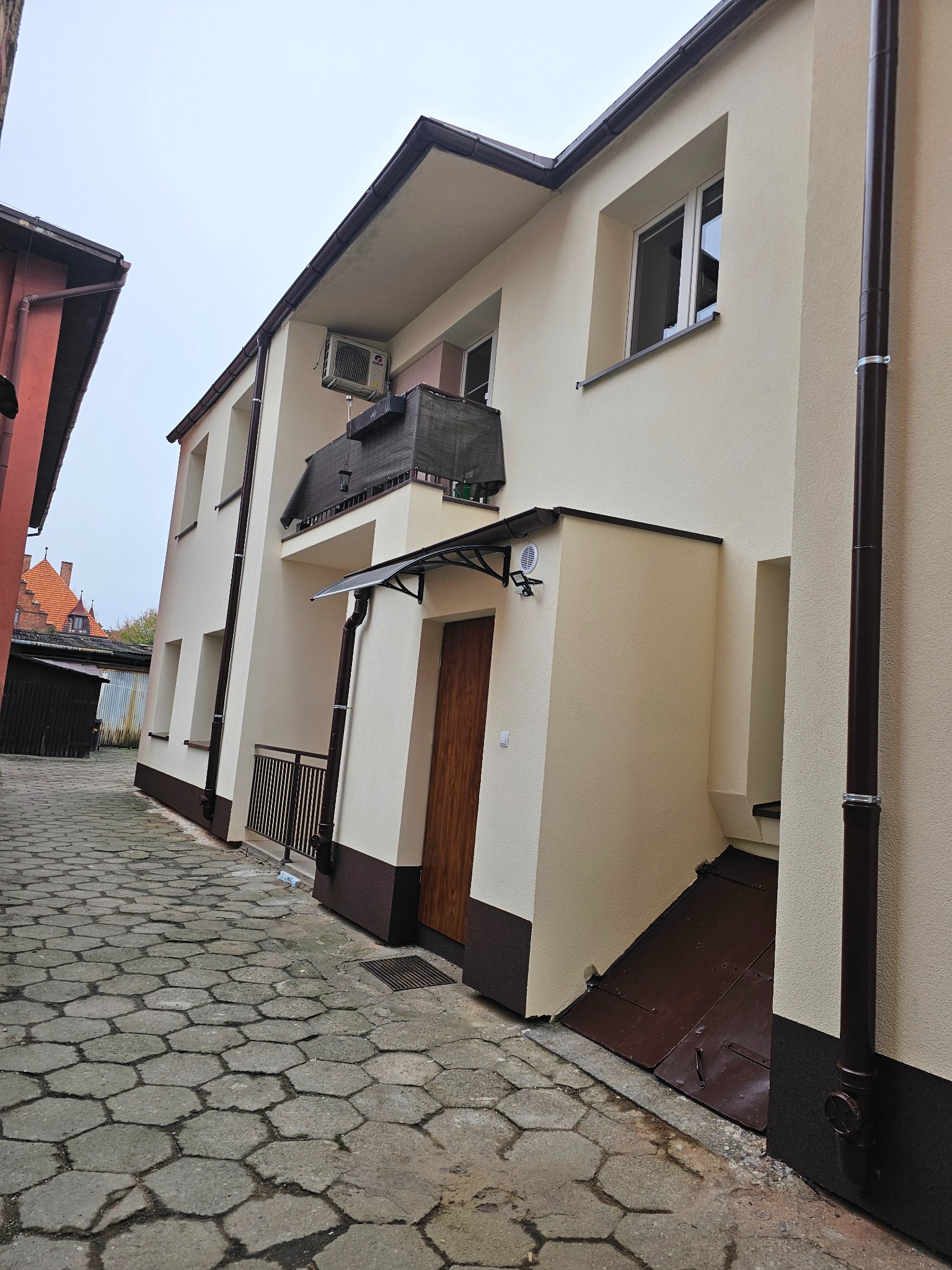 Jasny budynek z ocieploną elewacją, brązowe rynny i podmurówka, widok z wąskiej uliczki wyłożonej kostką.