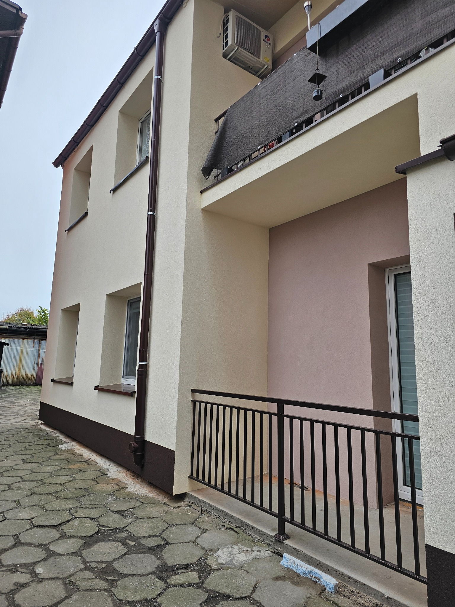 Dwupiętrowy budynek z beżową elewacją, brązowymi elementami wykończeniowymi i balkonem z czarną balustradą, widziany z dołu pod kątem.