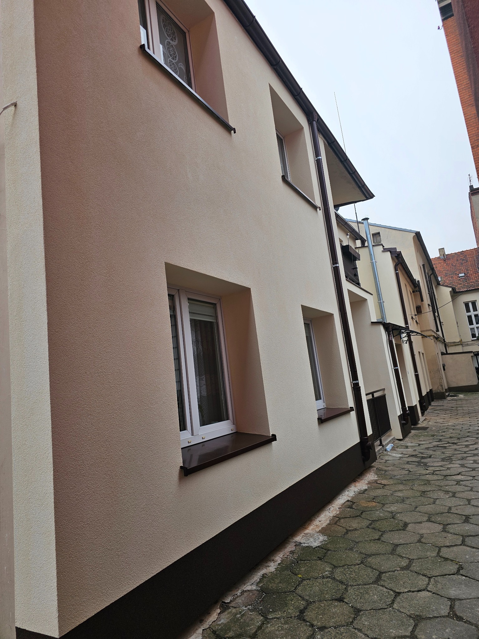 Elewacja budynku po renowacji z widoczną fakturą tynku i brązowymi parapetami, ujęcie z poziomu chodnika wyłożonego kostką.