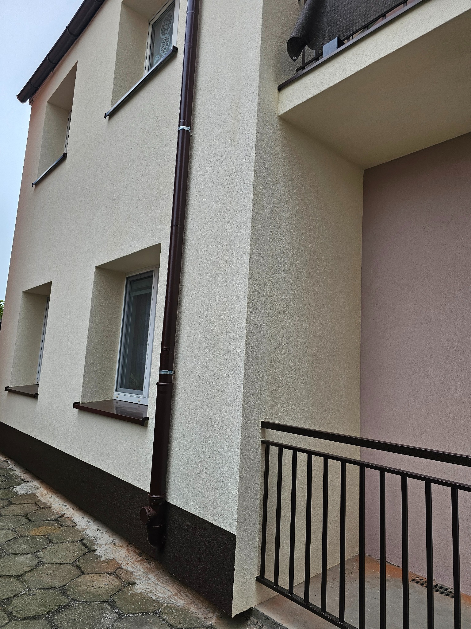 Dwukondygnacyjny budynek z jasną elewacją o ziarnistej fakturze, brązowymi rynnami i parapetami, widok z poziomu chodnika z betonowej kostki.