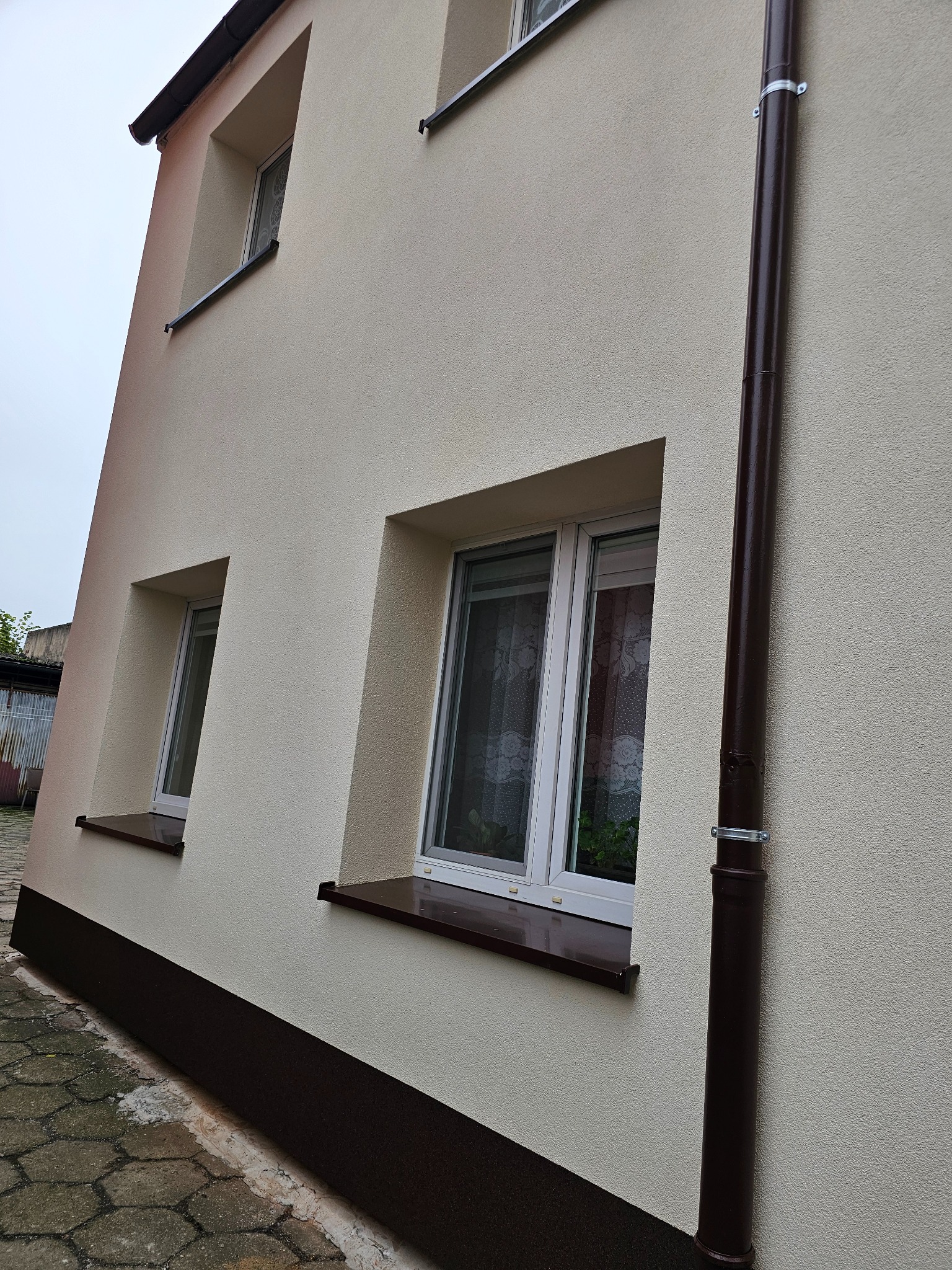 Elewacja budynku po renowacji z widocznymi oknami z brązowymi parapetami, rynną spustową i fragmentem chodnika z kostki.