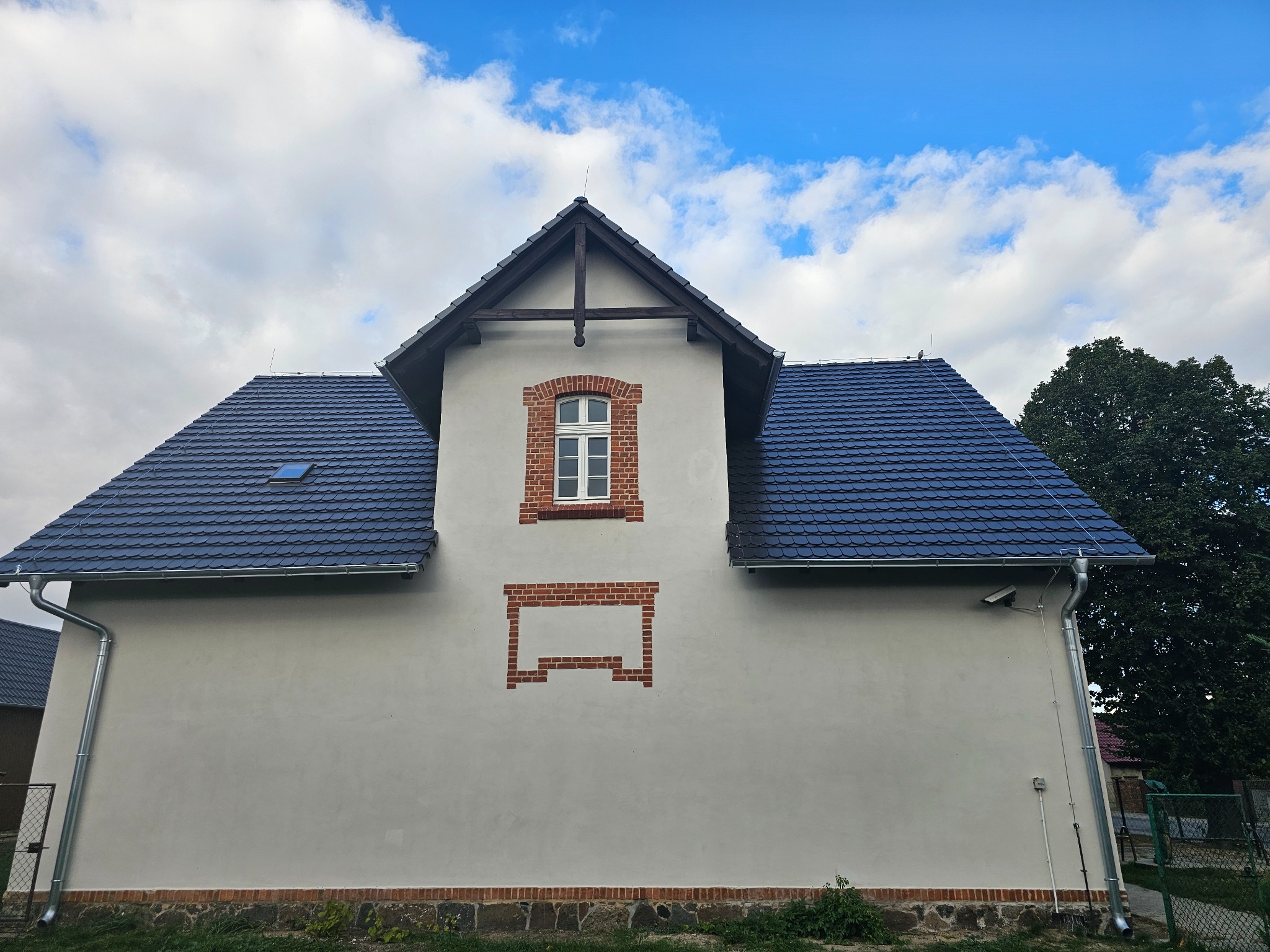 Elewacja budynku z nową, ciemną dachówką, jasnym tynkiem i dekoracyjnymi elementami z cegły wokół okna i w dolnej części ściany.