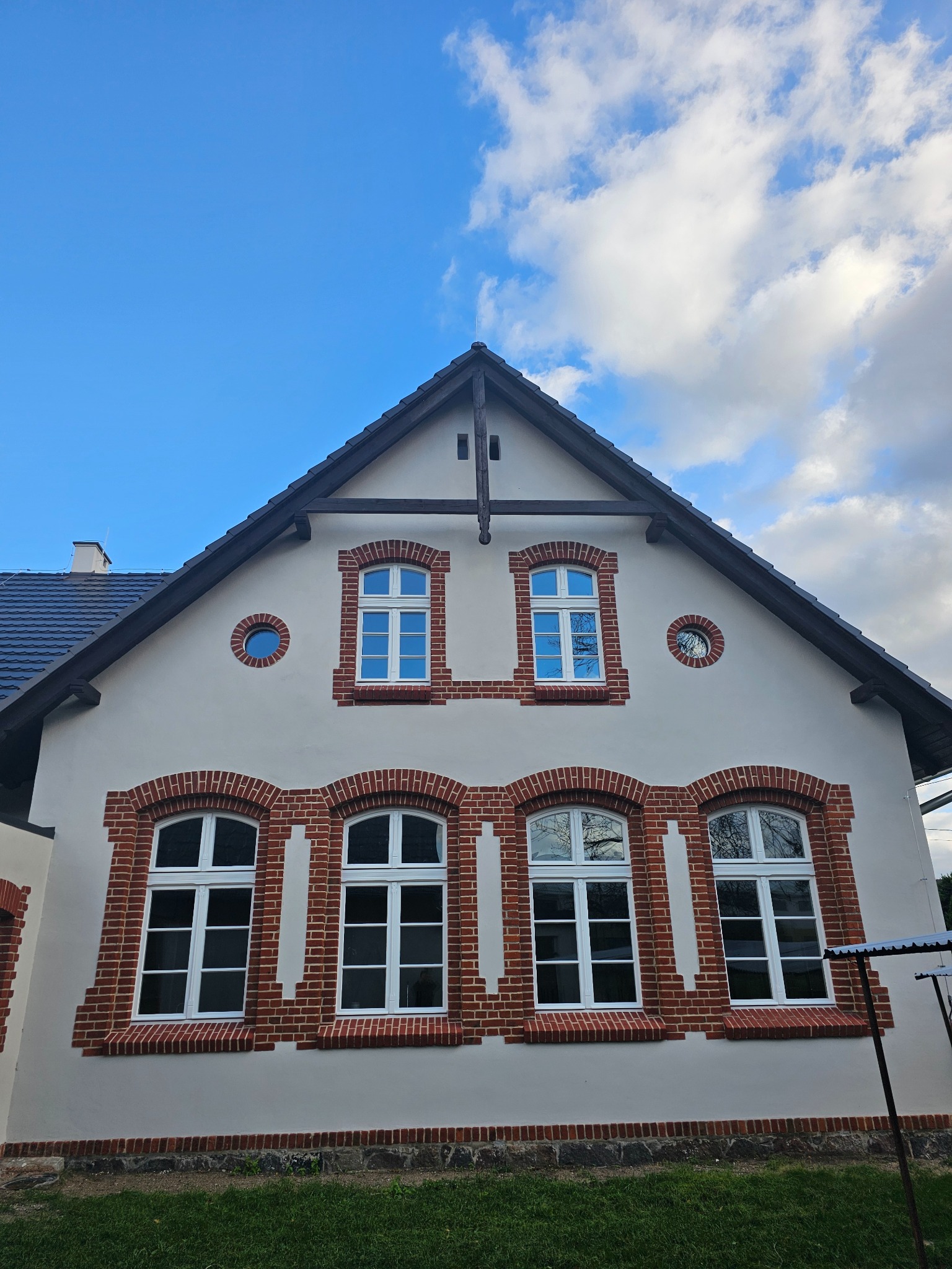 Odnowiona fasada domu z białym tynkiem i ceglaną obramówką okien, widoczne niebieskie niebo i fragment dachu.