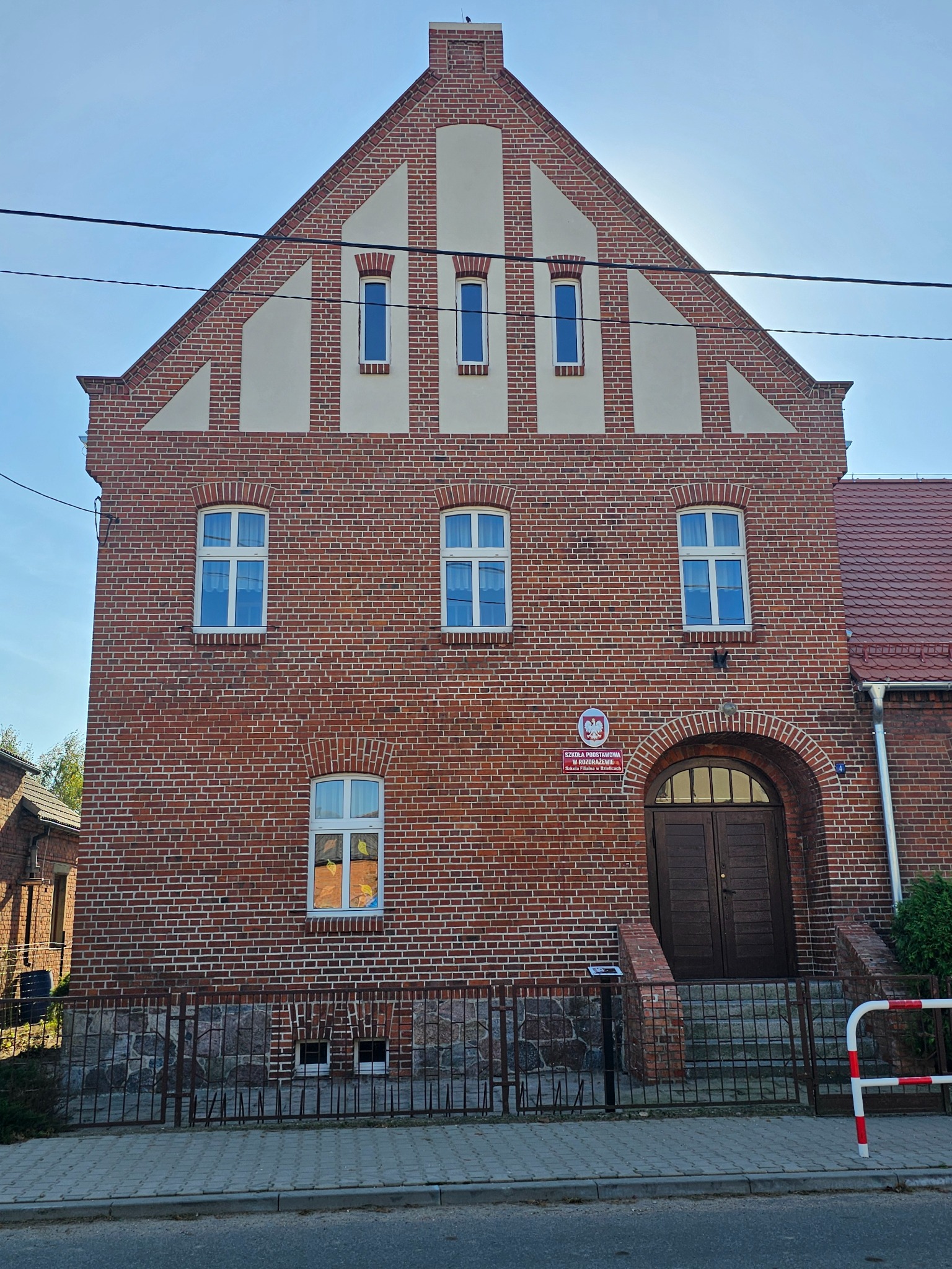 Dwupiętrowy budynek z czerwonej cegły z białymi akcentami na szczycie, widoczne okna z białymi ramami i drewniane drzwi wejściowe z łukowatym portalem, tablica informacyjna po prawej stronie drzwi.