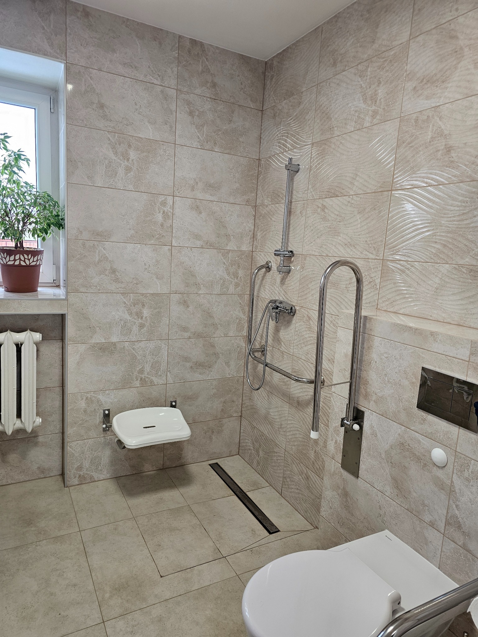 Beżowa łazienka z marmurkowymi płytkami, wyposażona w prysznic z siedziskiem dla niepełnosprawnych, poręcz, toaletę i grzejnik pod oknem z rośliną doniczkową.