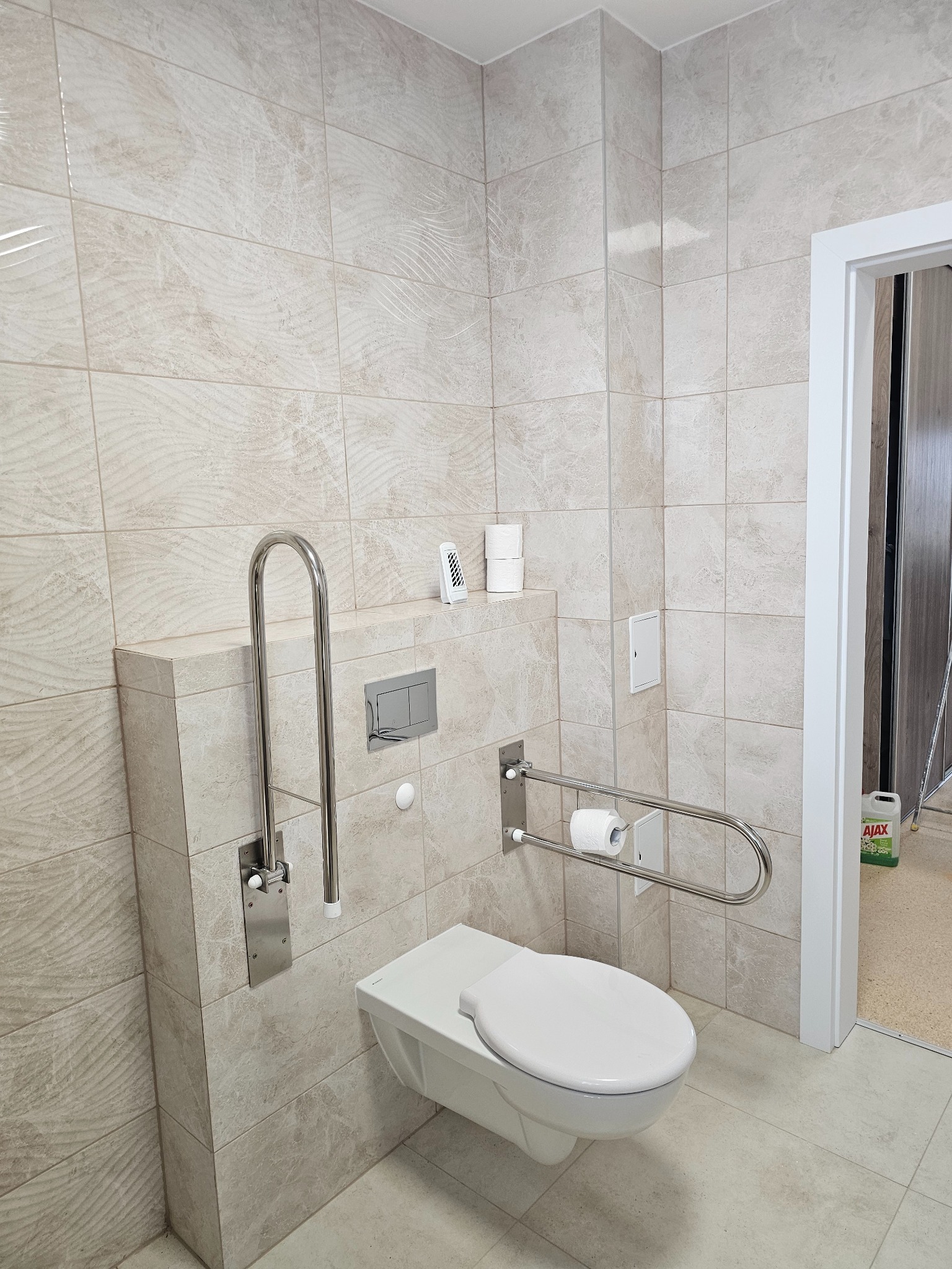 Beżowa łazienka z wiszącą toaletą, uchwytami dla niepełnosprawnych i płytkami imitującymi marmur, widoczny dozownik zapachu i zapas papieru toaletowego.