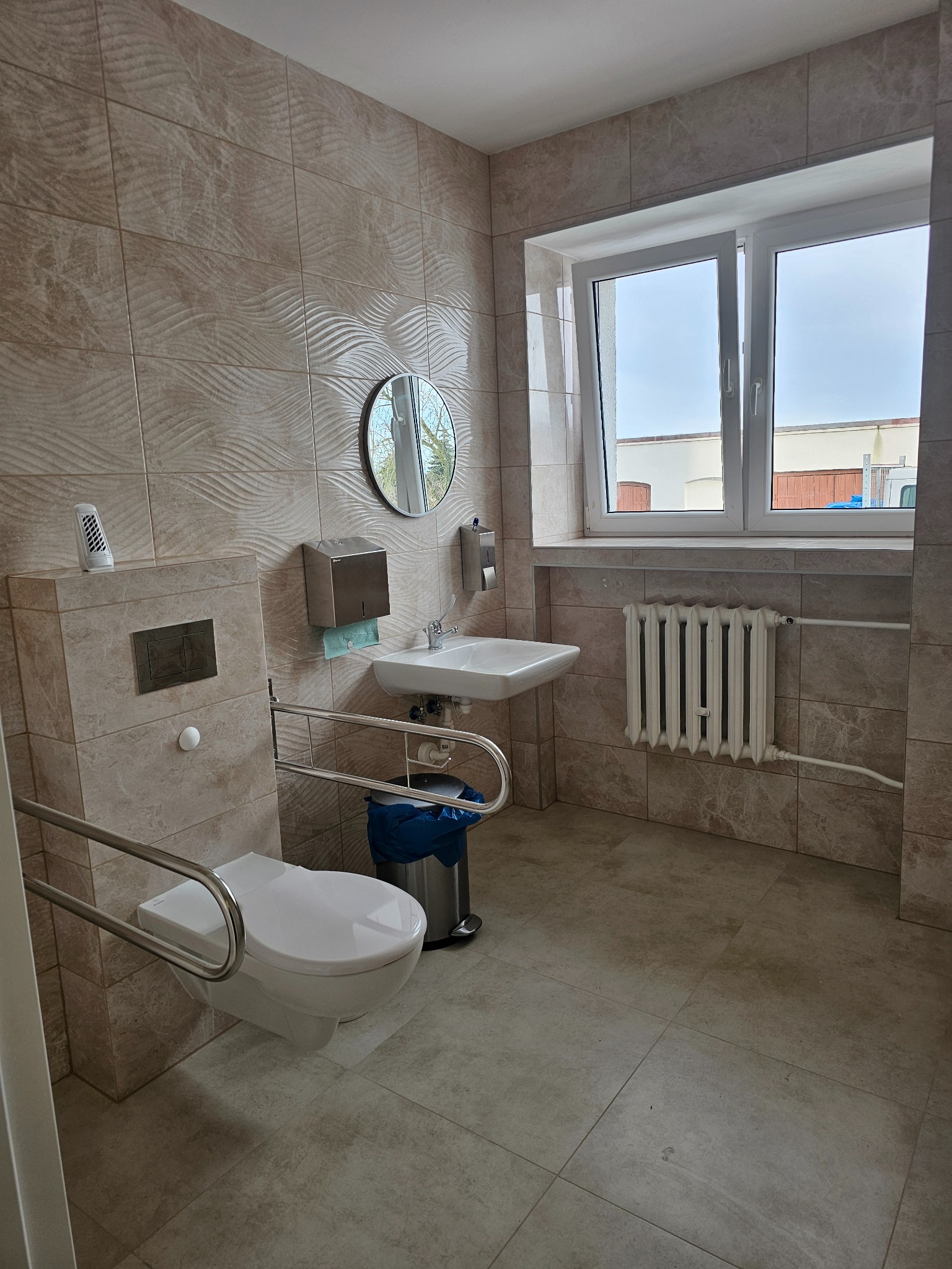 Umywalka, toaleta z uchwytami dla niepełnosprawnych oraz grzejnik w wykafelkowanej łazience z oknem. Na ścianach płytki z delikatnym wzorem fal. Widoczny kosz na śmieci i dozownik do mydła.