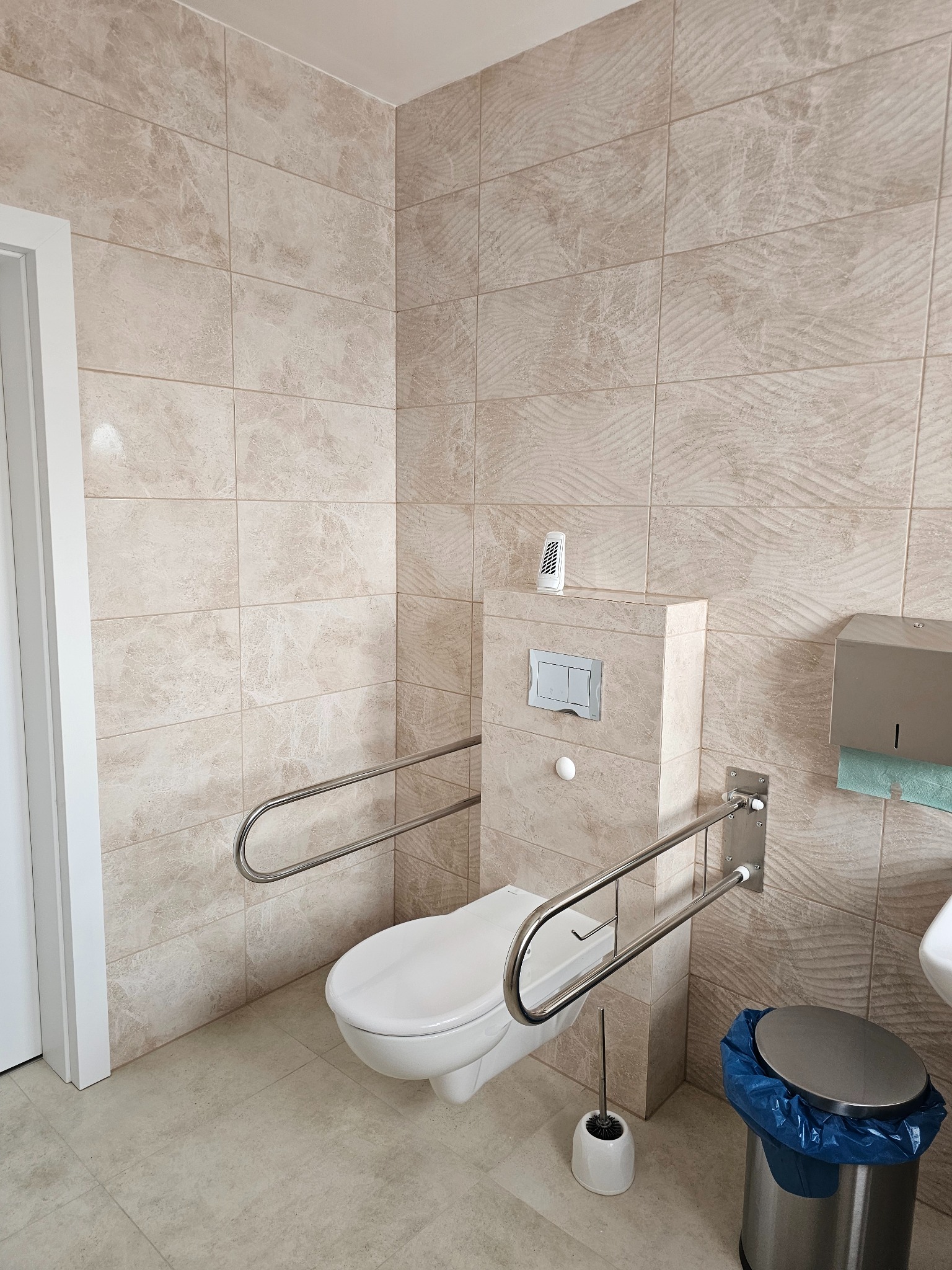 Beżowa łazienka z toaletą dla niepełnosprawnych, wyposażona w uchwyty, kosz na śmieci z niebieskim workiem i dozownik ręczników papierowych.