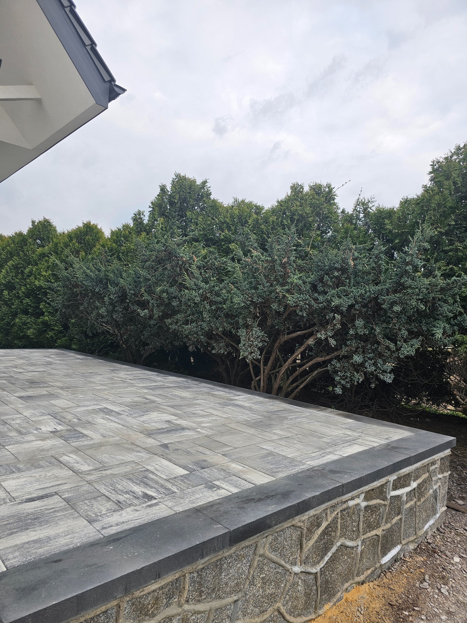 Wykończony taras z szarej kostki brukowej z obrzeżem, mur oporowy z kamienia polnego, tło z zielonych drzew iglastych i fragment dachu budynku z szarą blachodachówką.