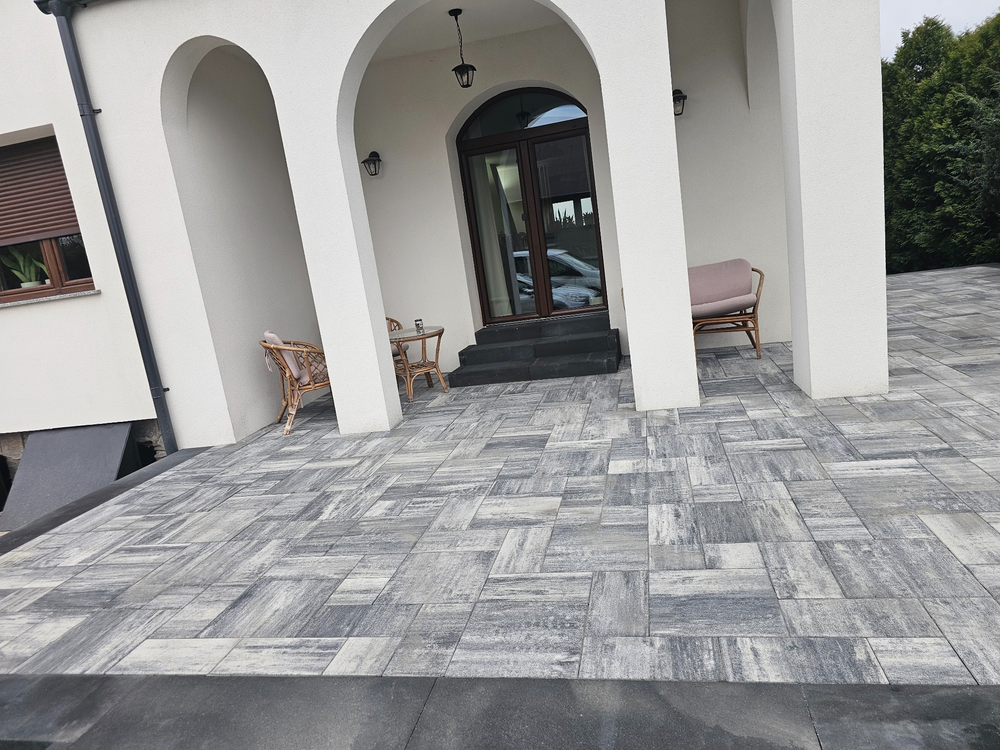 Ułożona szara kostka brukowa przed wejściem do domu z białymi kolumnami, widoczne meble ogrodowe i ciemne schody.