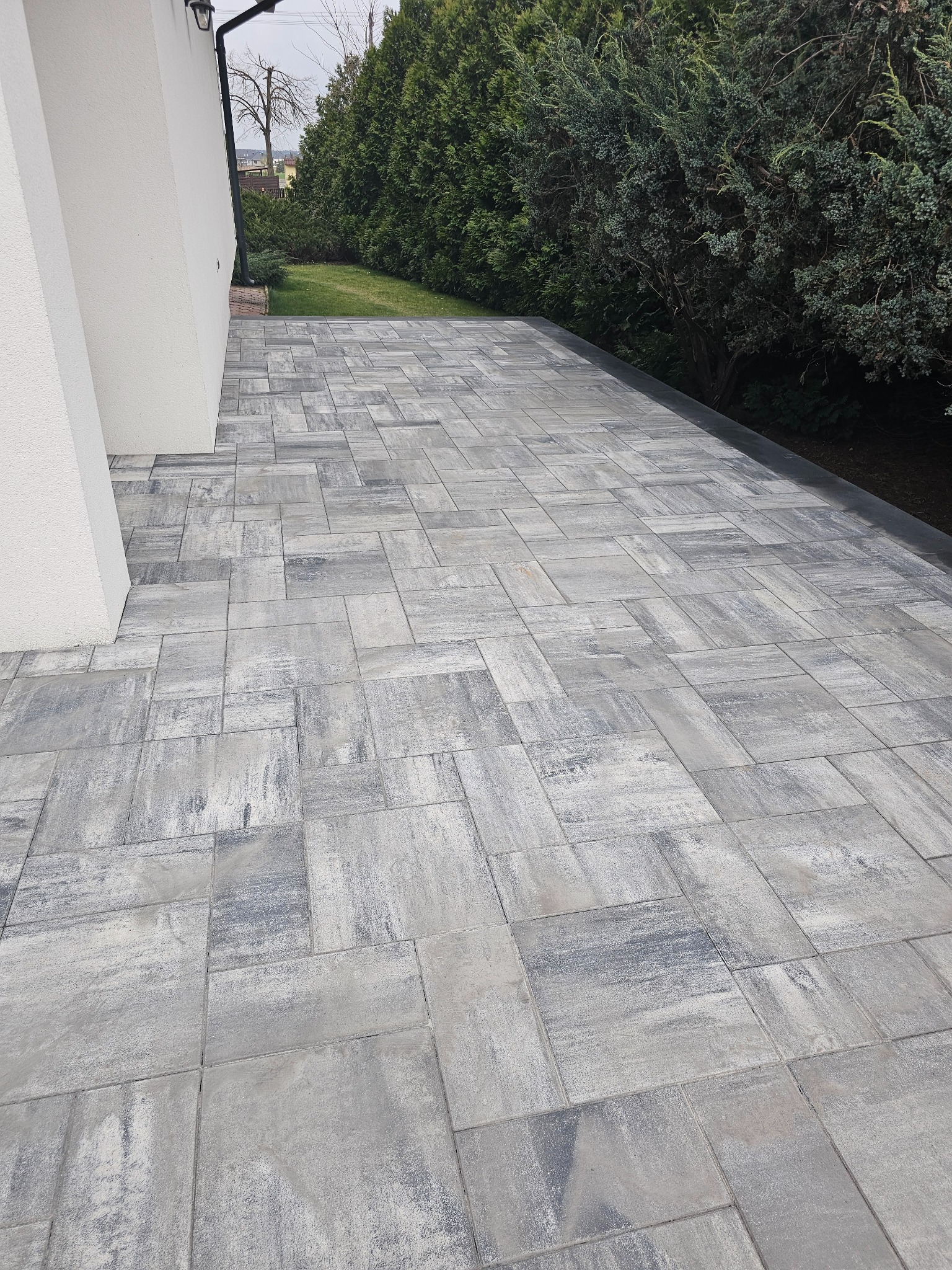 Ułożona szara kostka brukowa na tarasie przylegającym do białej ściany budynku, z zielonym żywopłotem w tle.