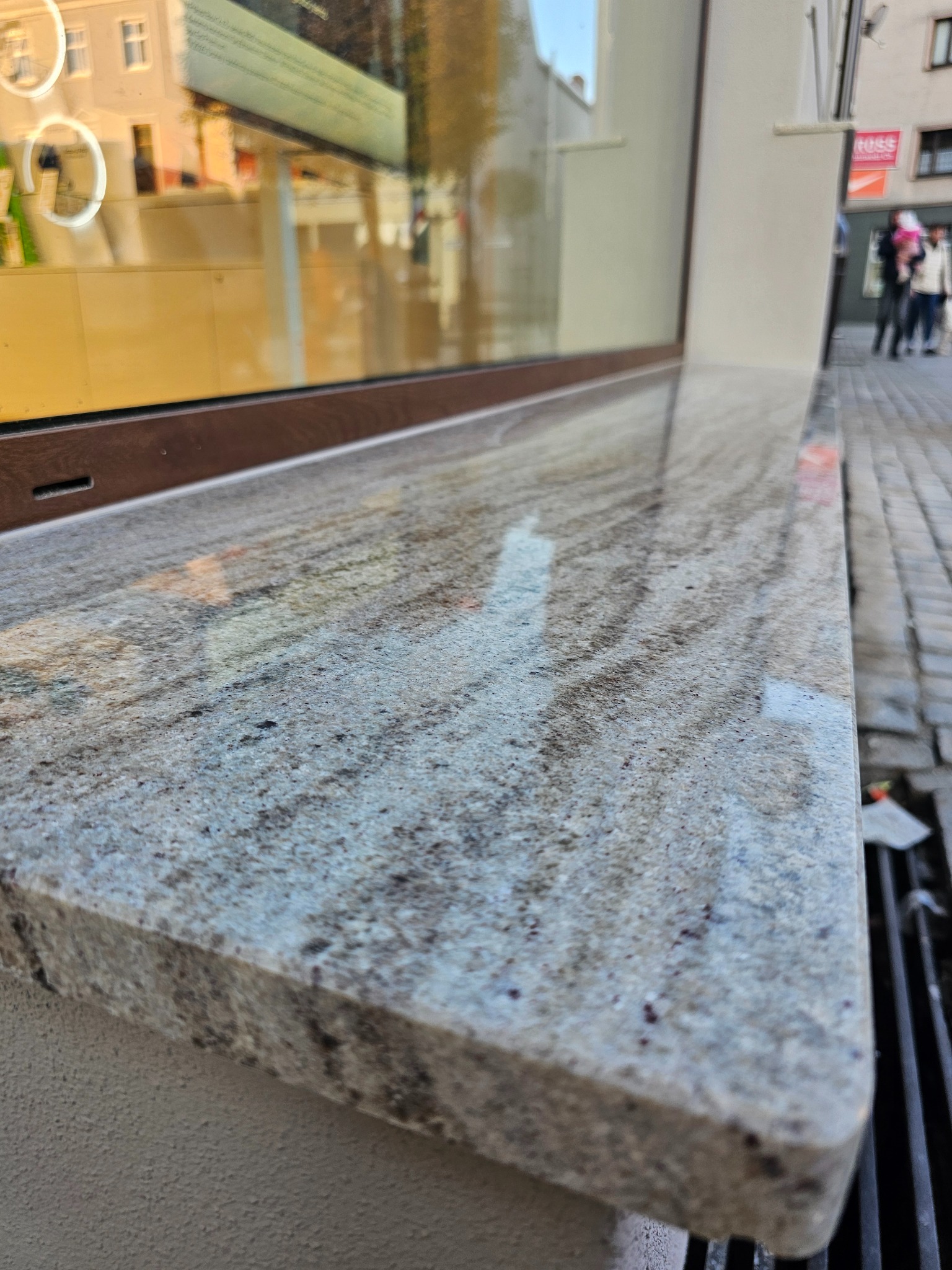 Granitowy parapet zewnętrzny z widoczną fakturą kamienia, zamontowany przy oknie budynku. W tle fragment chodnika i przechodnie.