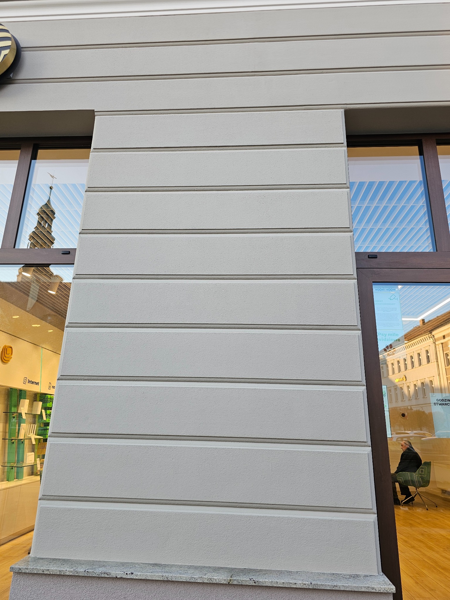 Szara elewacja budynku z poziomymi pasami dekoracyjnymi, widoczna część okna z odbiciem wieży kościoła i fragment wnętrza sklepu z siedzącą osobą.