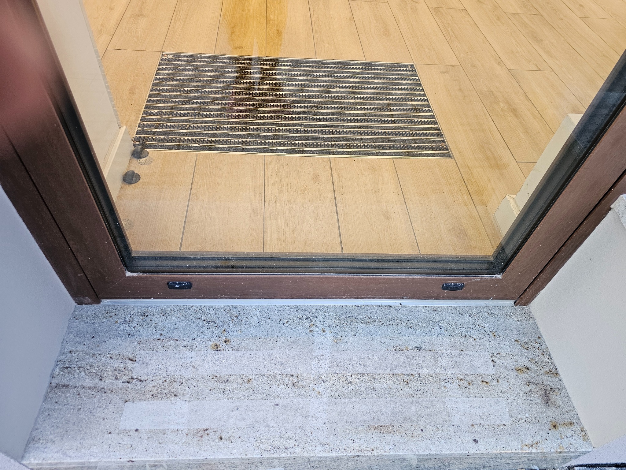 Nowoczesne wejście z drewnianą ramą drzwiową, jasną podłogą z płytek i wycieraczką wpuszczoną w podłogę. Widoczne detale wykończenia i kamienne stopnie.