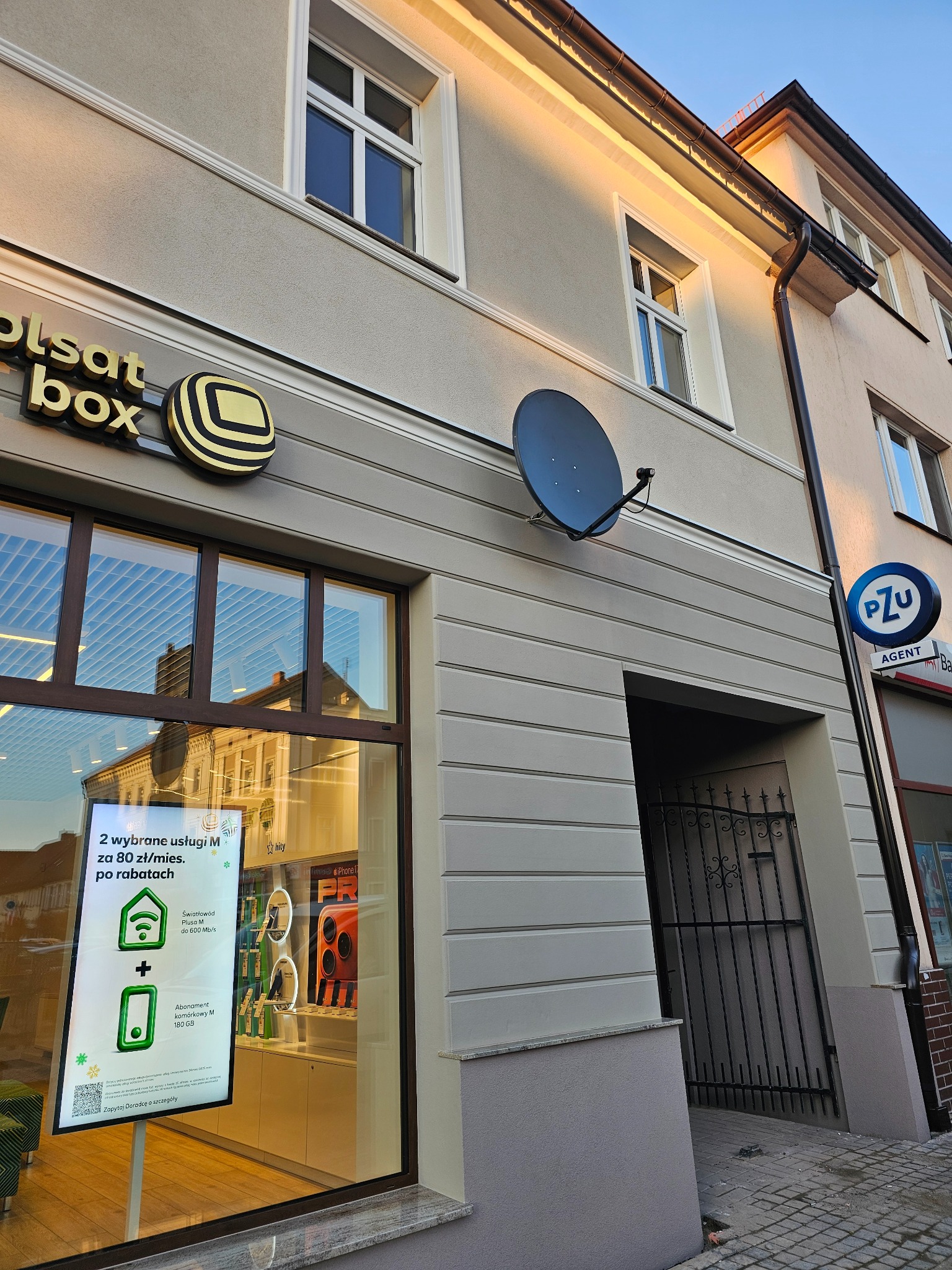 Elewacja budynku z logo Polsat Box i anteną satelitarną. Widoczne okna i szyld PZU. Budynek w jasnych barwach, oświetlony popołudniowym słońcem.
