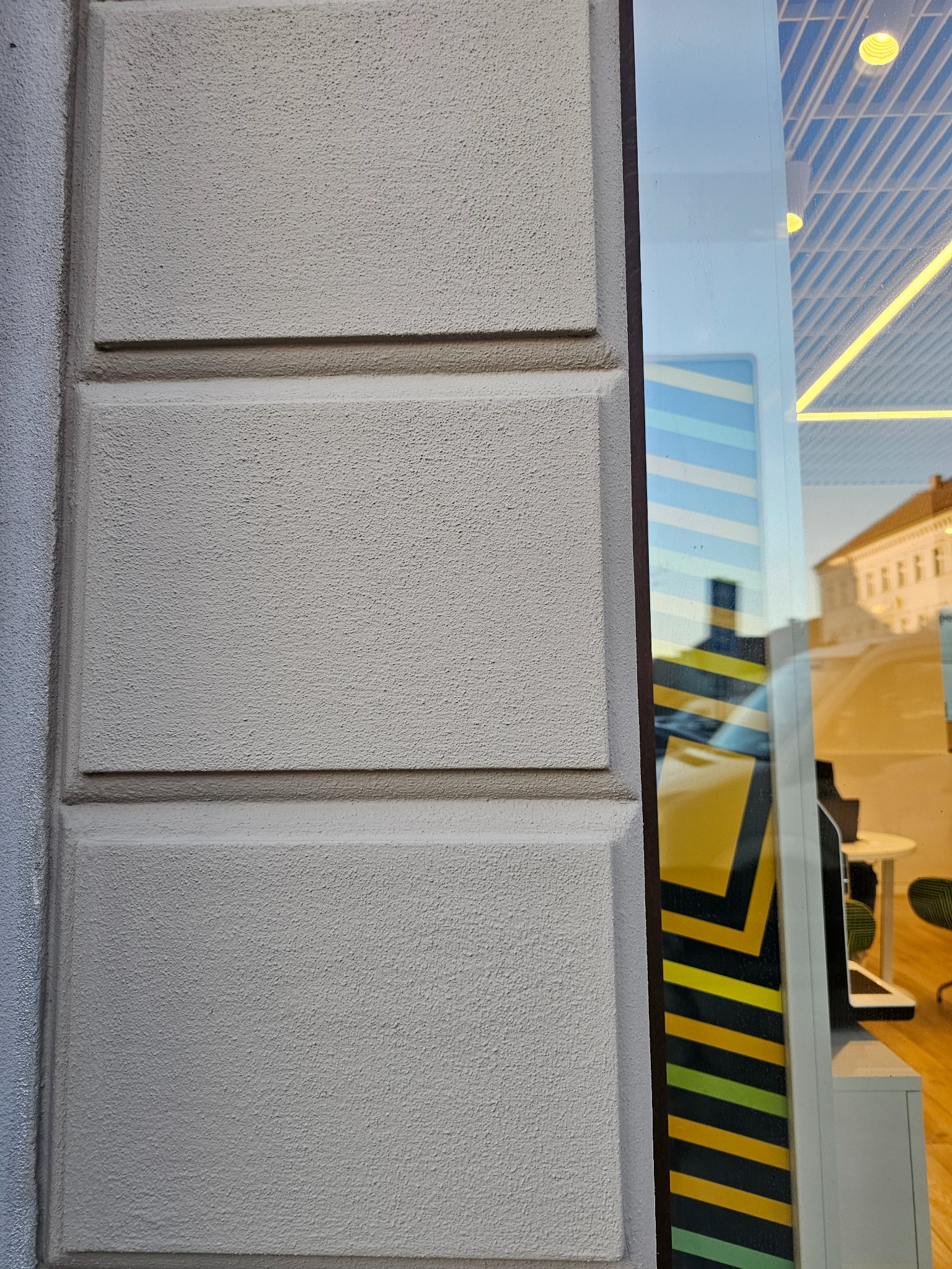 Fragment elewacji budynku z fakturą, widoczny obok okna wystawowego z odbiciami wnętrza i ulicy. Jasna, neutralna kolorystyka tynku.