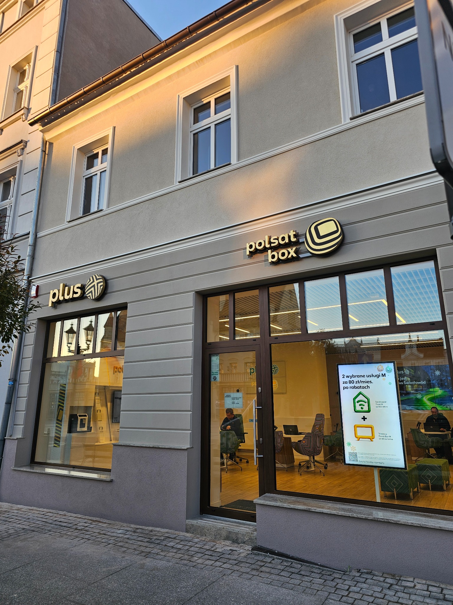 Fasada budynku z szyldami 'plus' i 'polsat box' nad wejściem. Widoczne okna i drzwi wejściowe. Wnętrze biura widoczne przez szybę.