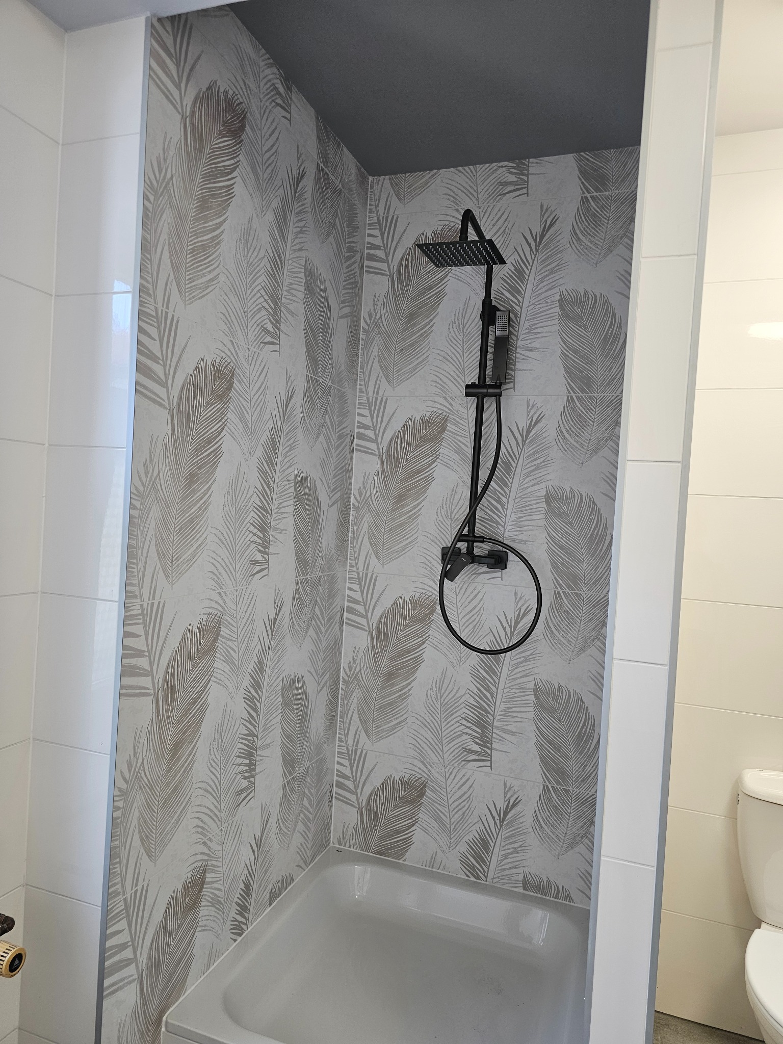 Wykończona łazienka z kabiną prysznicową i wanną. Ściany wyłożone płytkami z motywem piór, czarna armatura. Widoczny fragment toalety.