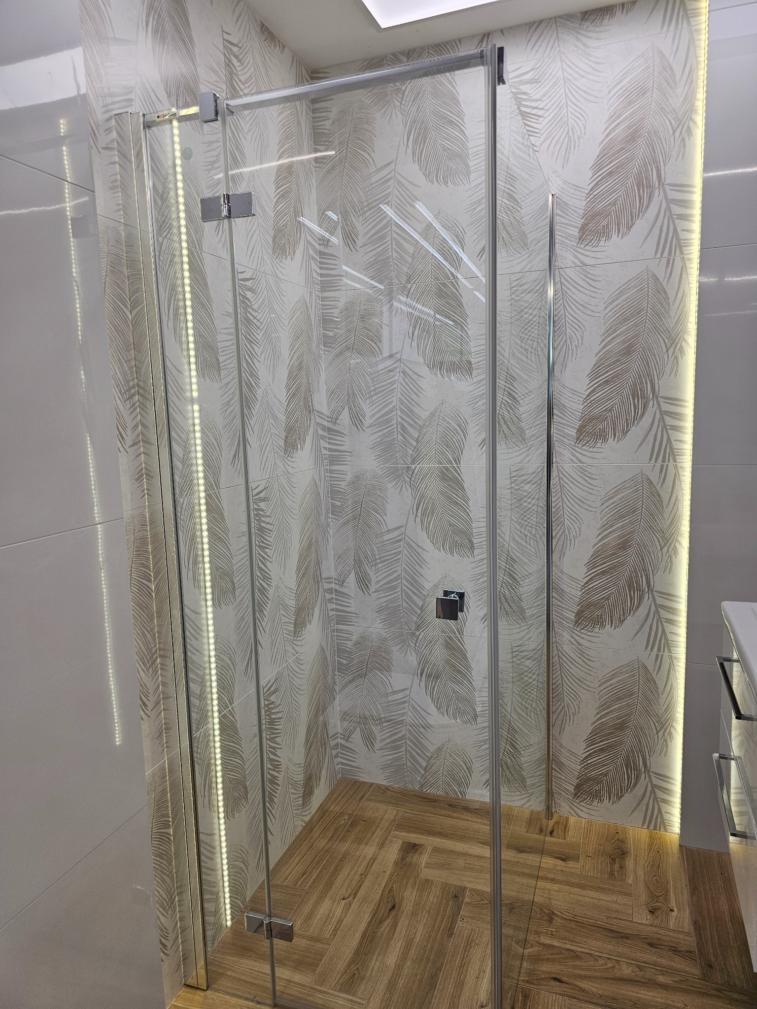 Szklana kabina prysznicowa z chromowanymi okuciami, na tle płytek z motywem liści i drewnopodobnej podłogi ułożonej w jodełkę. Oświetlenie LED podkreśla krawędzie.