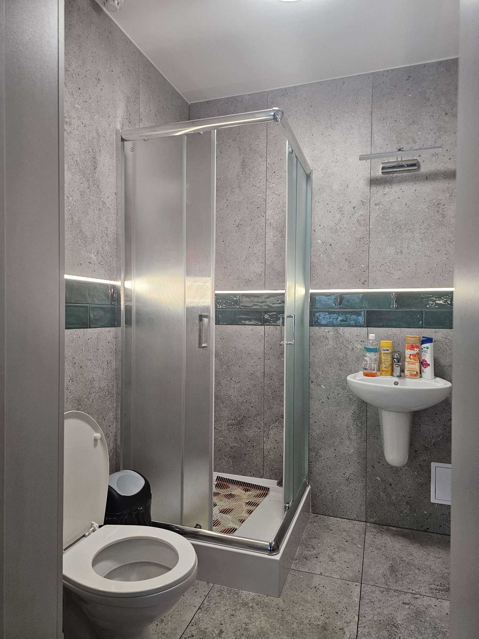 Szara łazienka z kabiną prysznicową, umywalką i toaletą. Ściany wyłożone płytkami imitującymi beton, z dekoracyjnym pasem zielonych płytek. Nowoczesne oświetlenie LED.