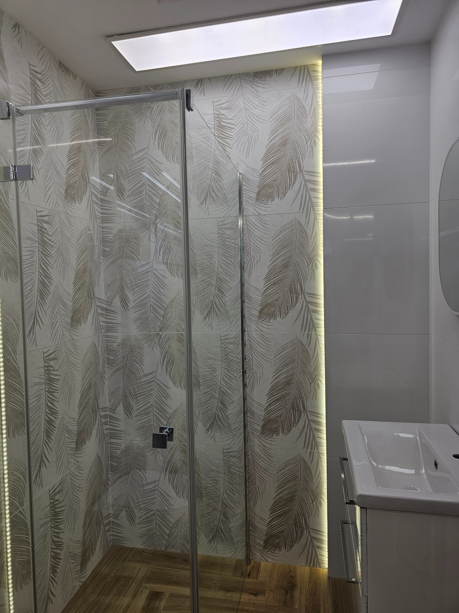 Nowoczesna kabina prysznicowa z deseniem liści na płytkach w Ostrowie Wielkopolskim. Biała umywalka i podświetlenie LED dodają elegancji.