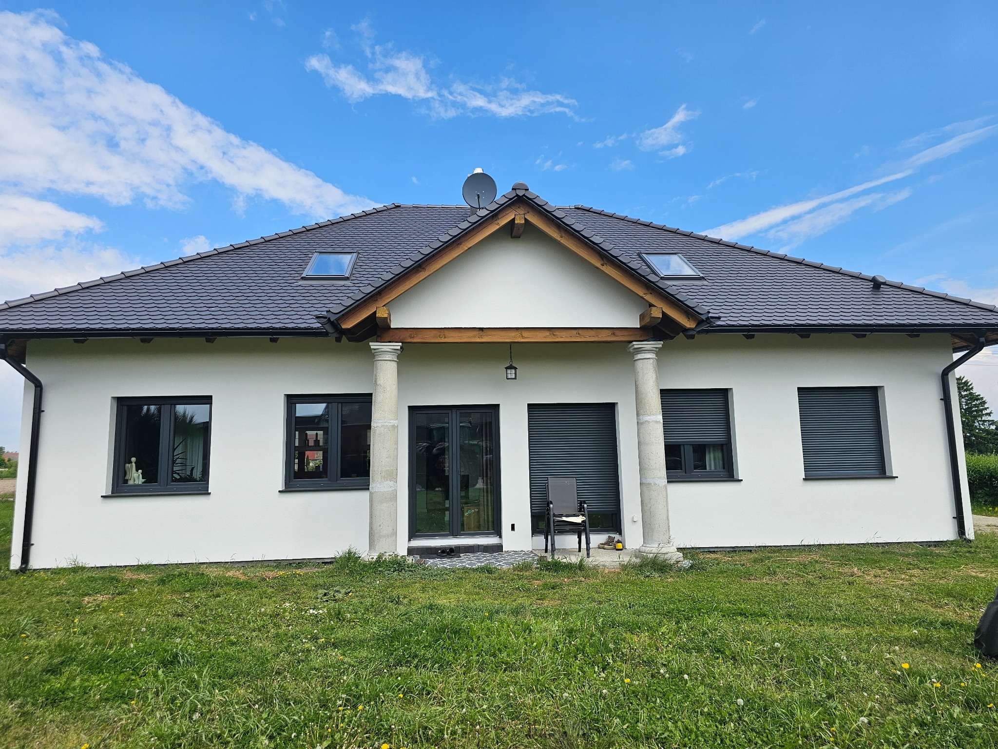 Biały dom jednorodzinny z ciemną dachówką i kolumnami przy wejściu, na tle błękitnego nieba z chmurami. Widoczne okna z szarymi ramami i roletami.