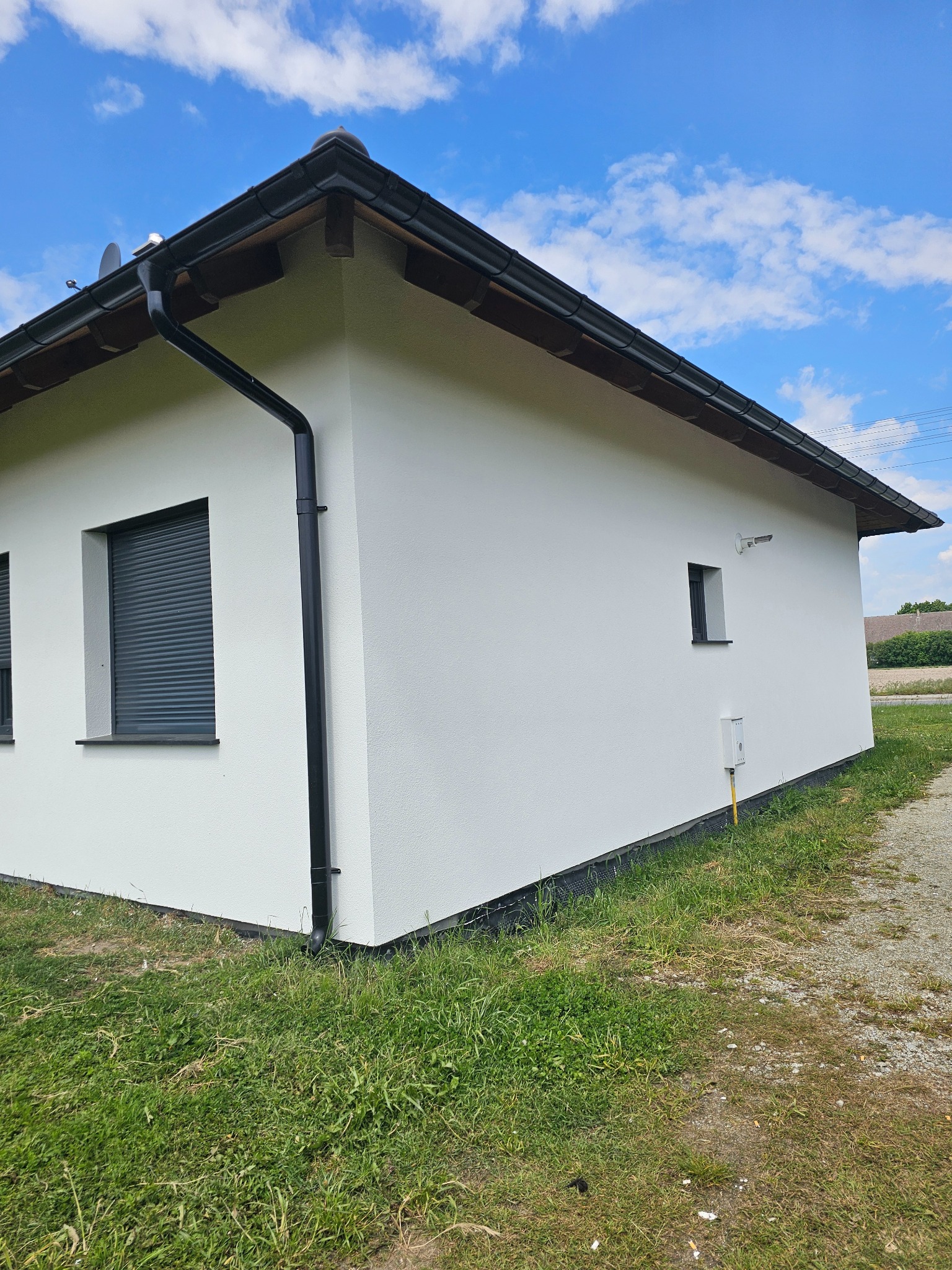 Boczny widok otynkowanego na biało domu z czarnymi rynnami i roletami okiennymi na tle błękitnego nieba z chmurami. Widoczny fragment trawnika i ścieżki.