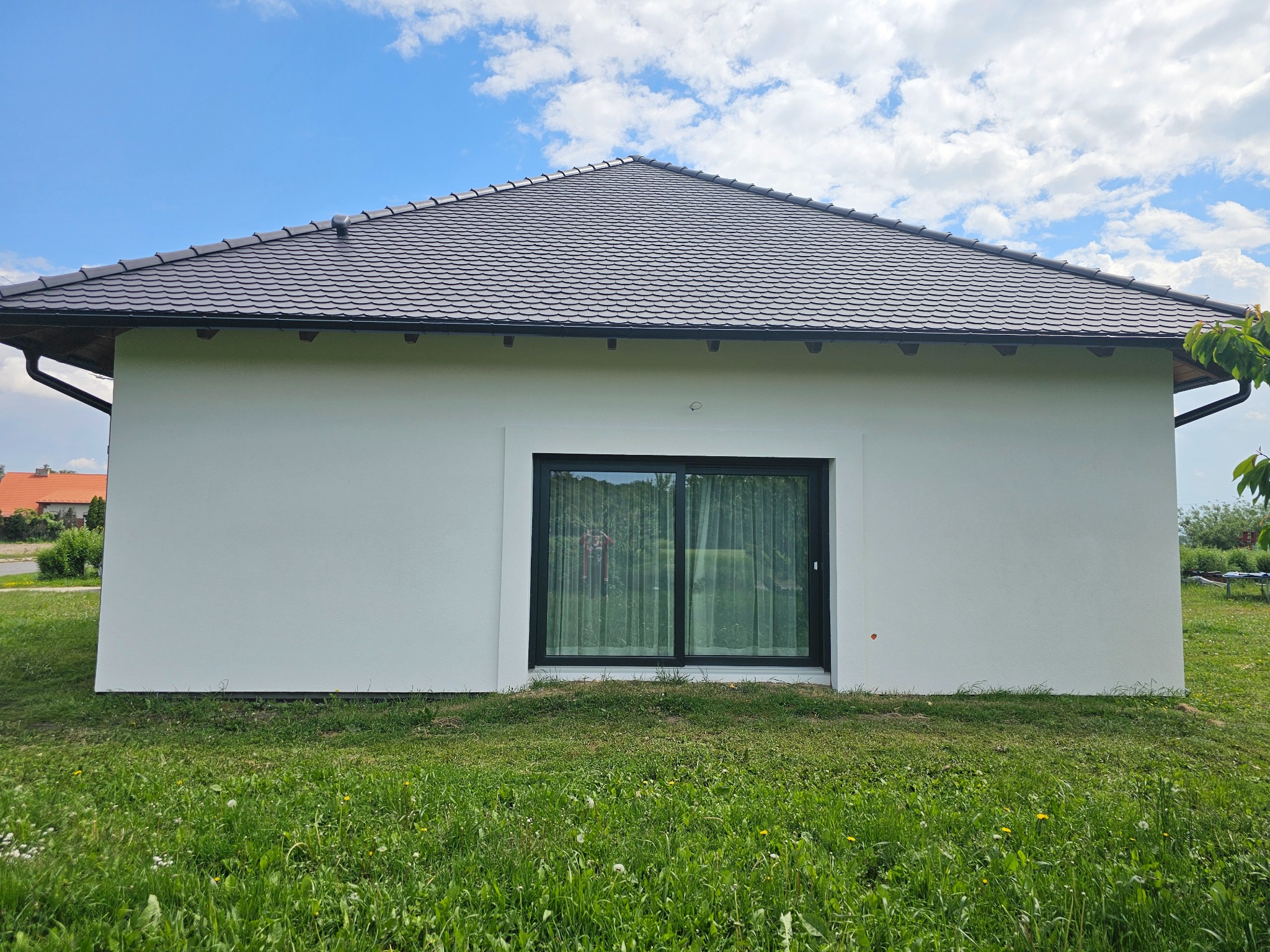 Boczny widok domu z białymi ścianami i ciemnym dachem w kształcie litery A, okno z czarną ramą, trawnik z przodu, błękitne niebo z chmurami w tle.