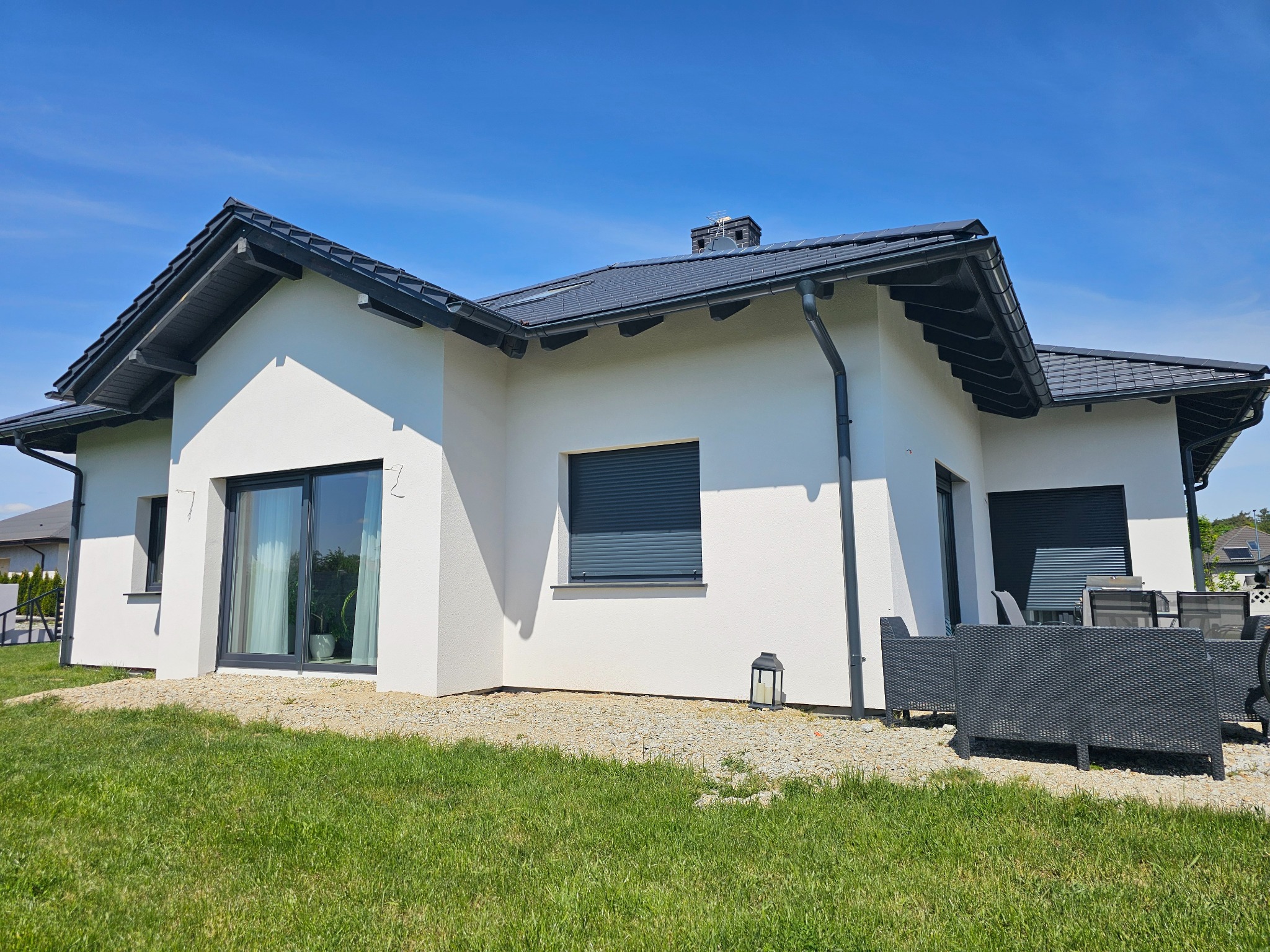 Biały dom jednorodzinny z ciemną dachówką i stolarką okienną, widok z ogrodu w słoneczny dzień. Minimalistyczny design, nowoczesna architektura.