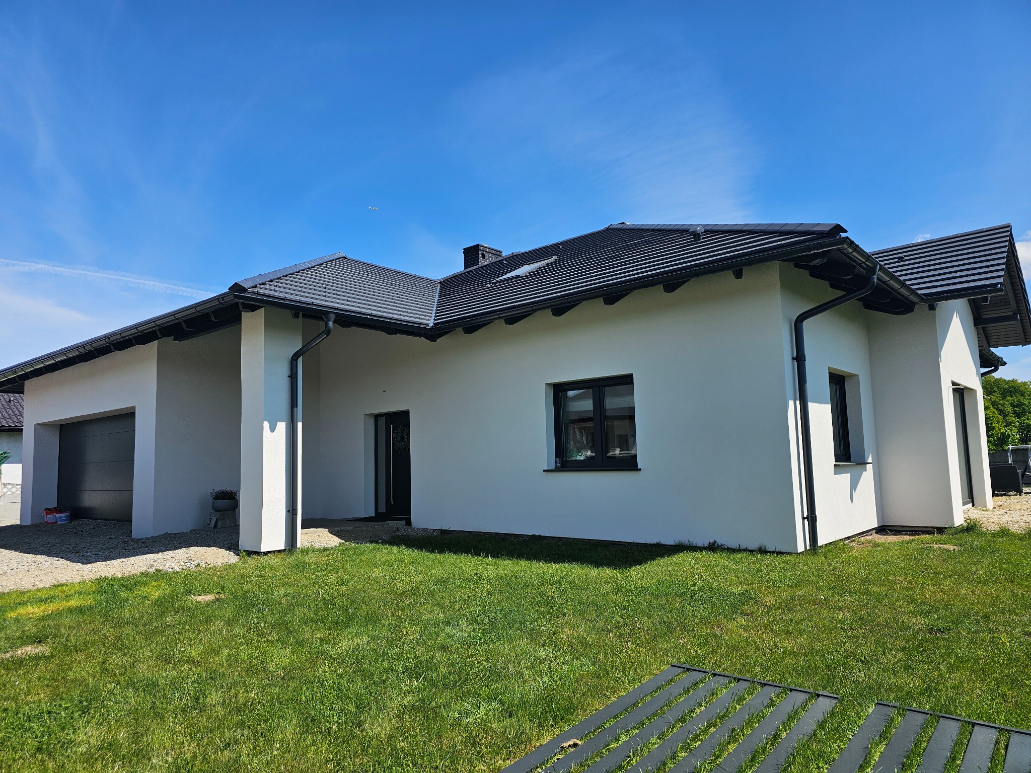 Nowoczesny dom jednorodzinny z białymi ścianami i ciemną dachówką, widok z ogrodu w słoneczny dzień. Widoczne detale wykończenia elewacji i zagospodarowania terenu.