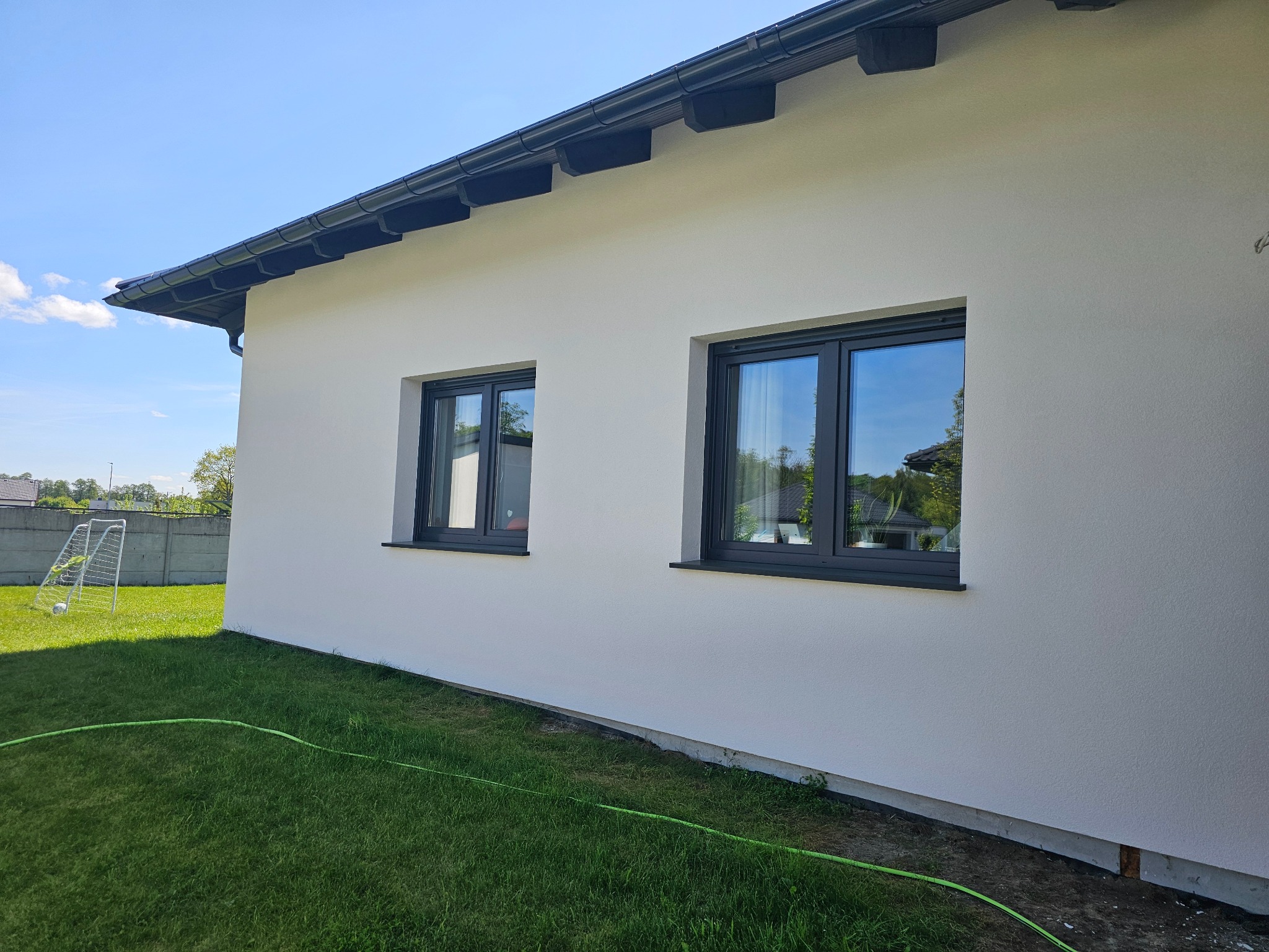 Biała elewacja domu jednorodzinnego z dwoma oknami w grafitowych ramach. Na trawniku leży zielony wąż ogrodowy, a w tle widoczna bramka do piłki nożnej.