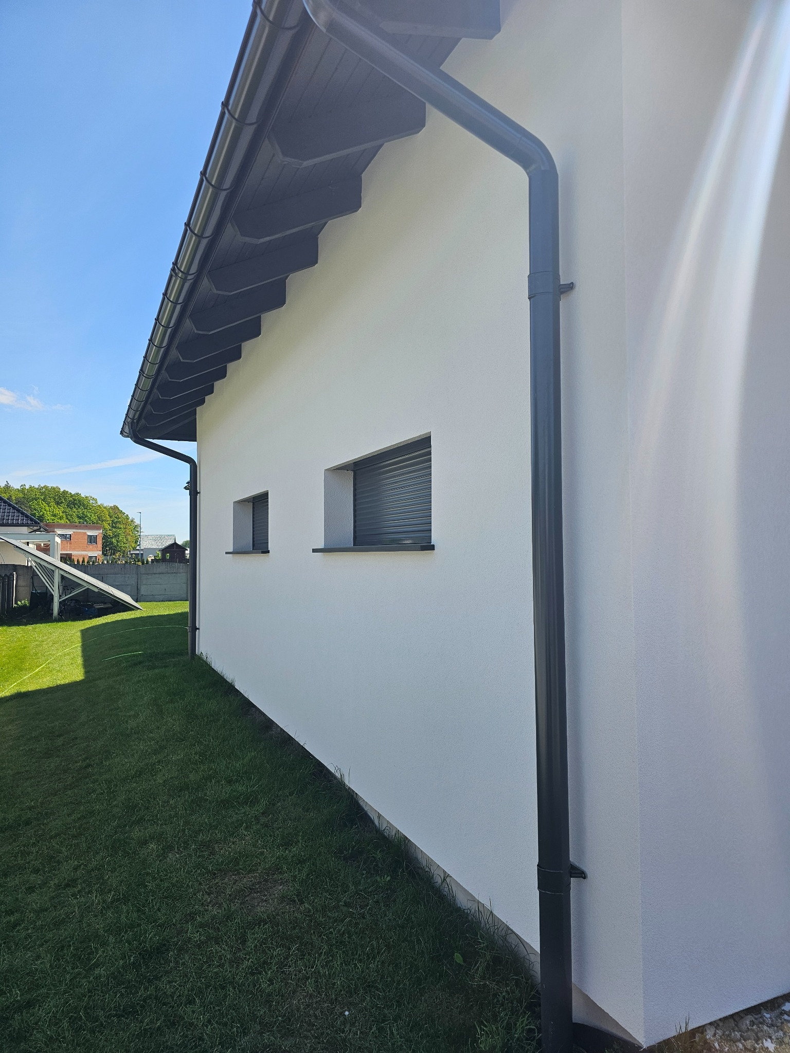 Biała elewacja budynku jednorodzinnego z szarymi oknami i rynną. Widok z zewnątrz na trawnik i panele słoneczne w tle. Słoneczny dzień.