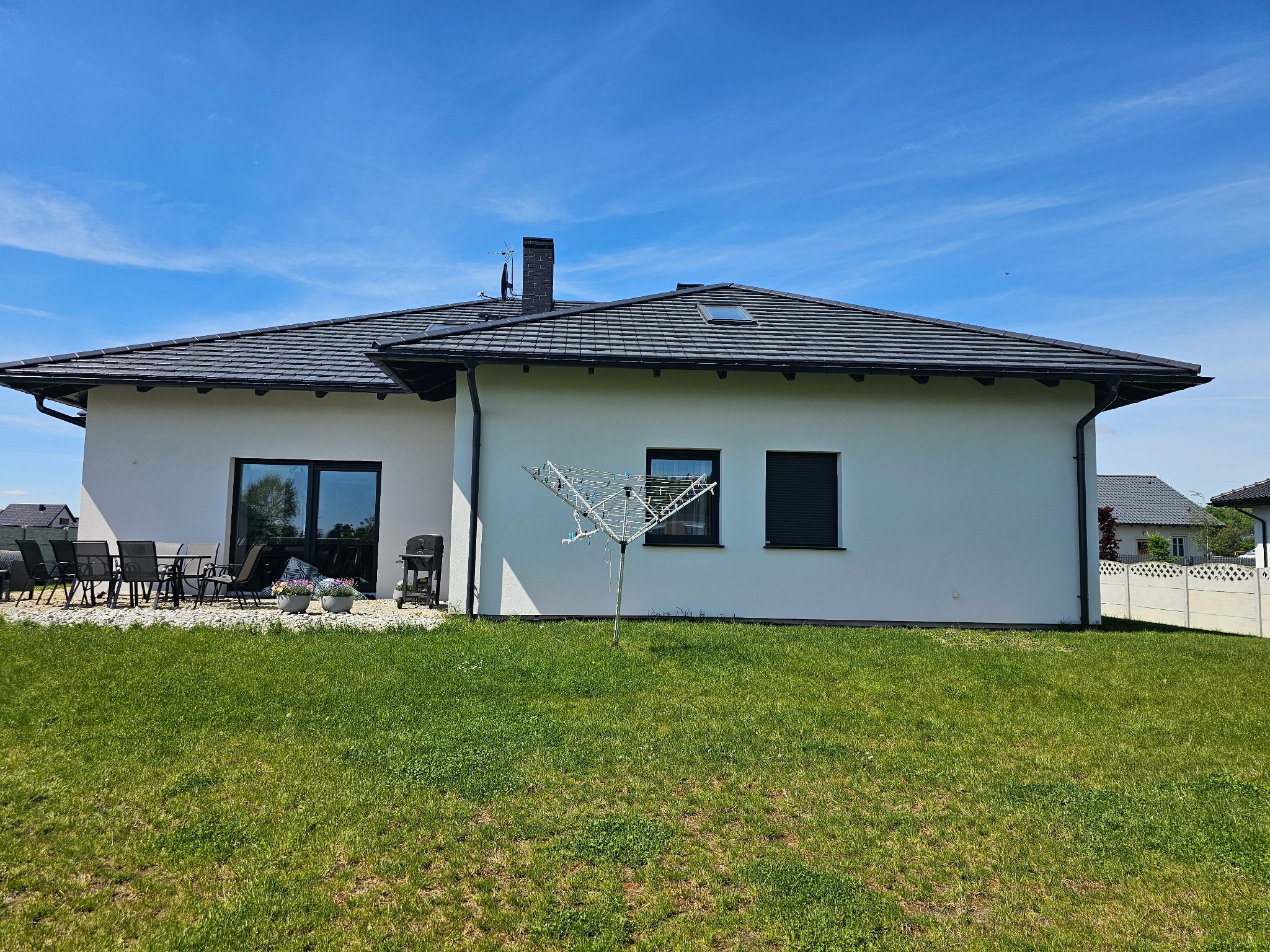 Boczny widok domu jednorodzinnego z białymi ścianami i ciemną dachówką, na pierwszym planie trawnik. Widoczne okna z roletami zewnętrznymi i taras z meblami ogrodowymi.