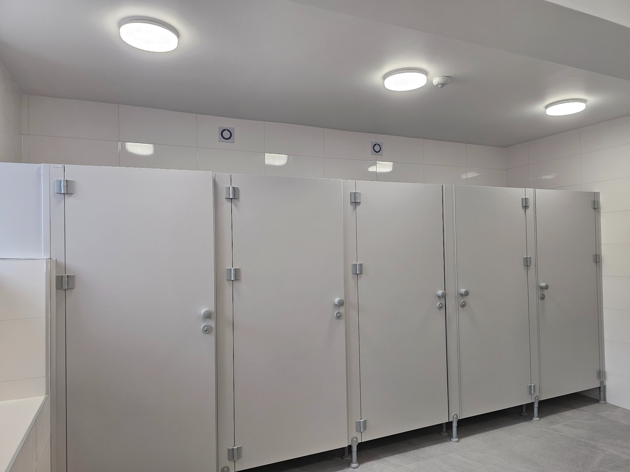 Rząd sześciu białych kabin toaletowych w nowoczesnej łazience publicznej, z białymi płytkami ściennymi, szarą podłogą i okrągłymi lampami sufitowymi LED.
