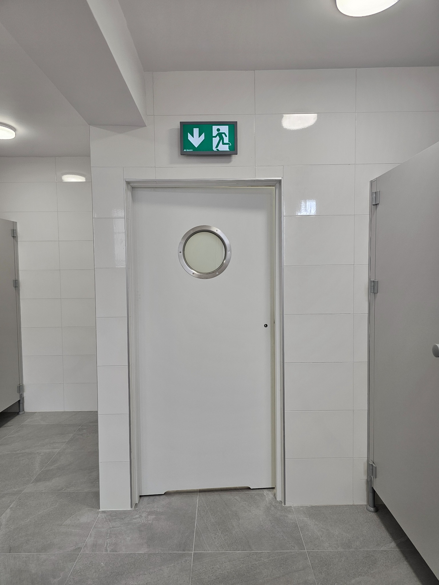 Nowoczesne wnętrze z białymi, prostokątnymi płytkami ściennymi i szarymi płytkami podłogowymi. Białe drzwi z okrągłym oknem, nad którymi zielony znak wyjścia ewakuacyjnego.